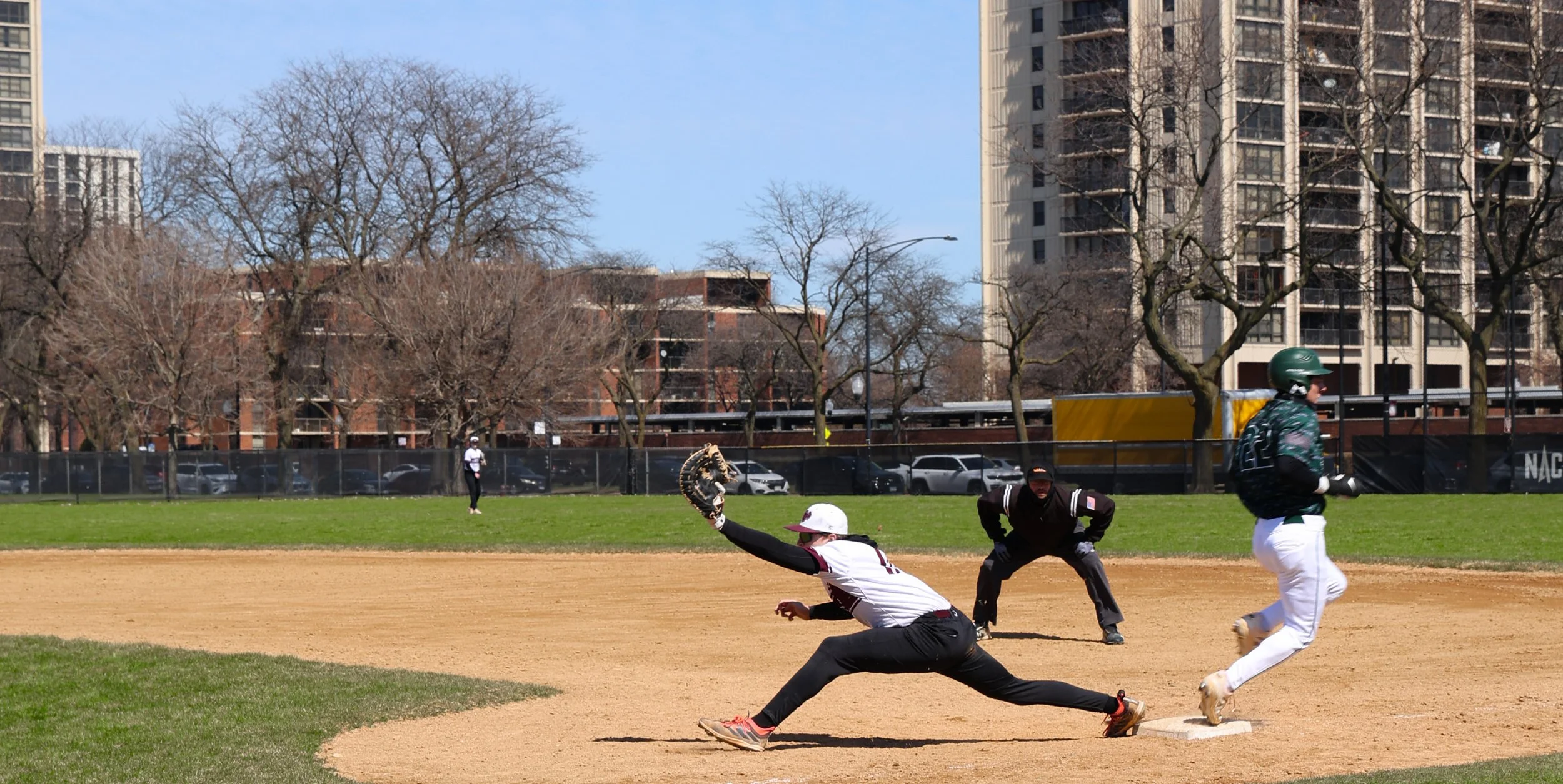 Baseball game in progress with players on the field, including a sliding player and a runner near the base, in an urban park setting with trees and tall buildings in the background.