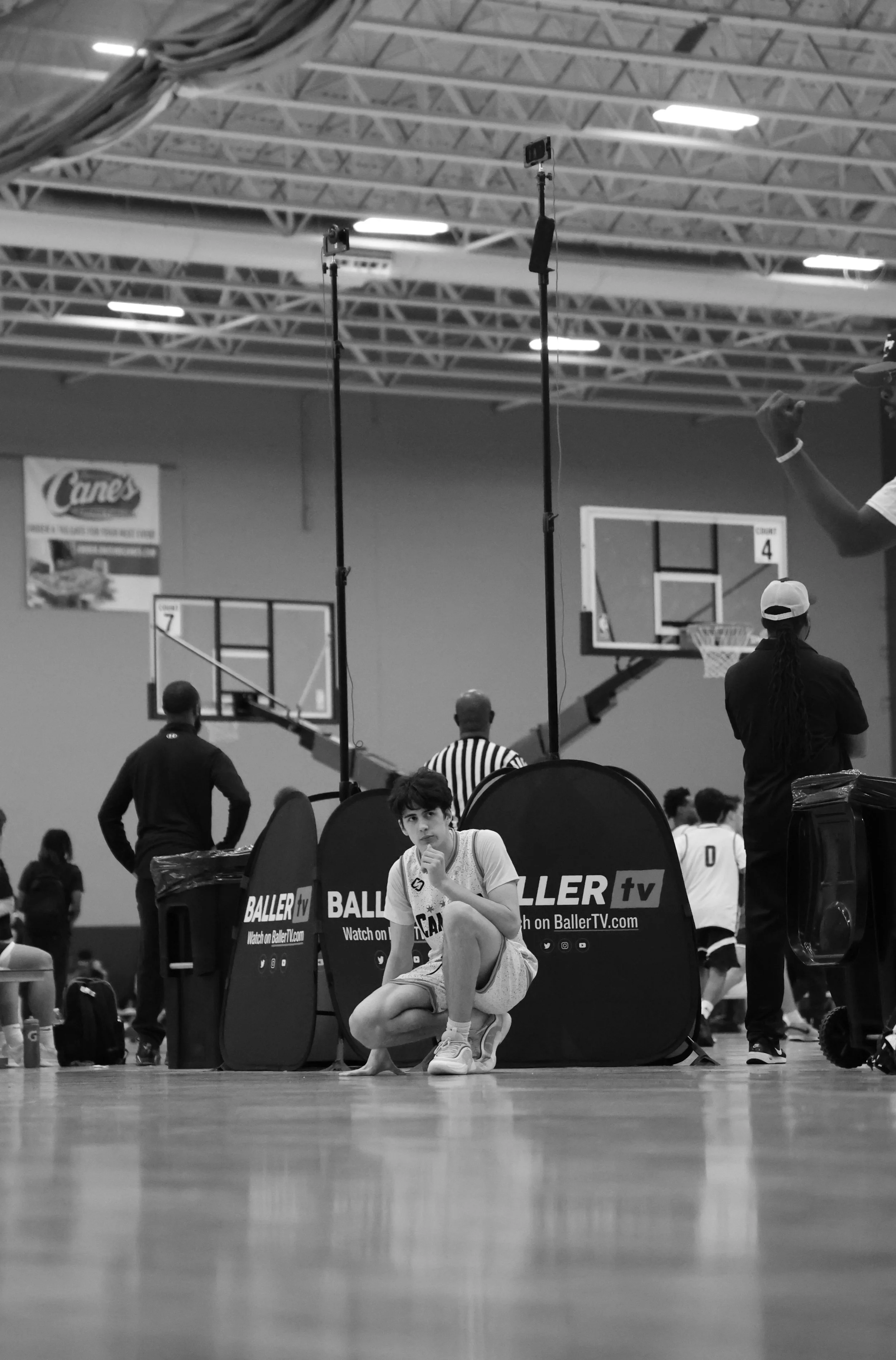 A young basketball player squats on the court in front of a black banner with the words "BALLER tv" and the website "BallerrTV.com." The player is wearing a white basketball uniform and has one hand resting on her knee while the other touches her chin. In the background, there are spectators, referees, and basketball hoops inside a gymnasium.