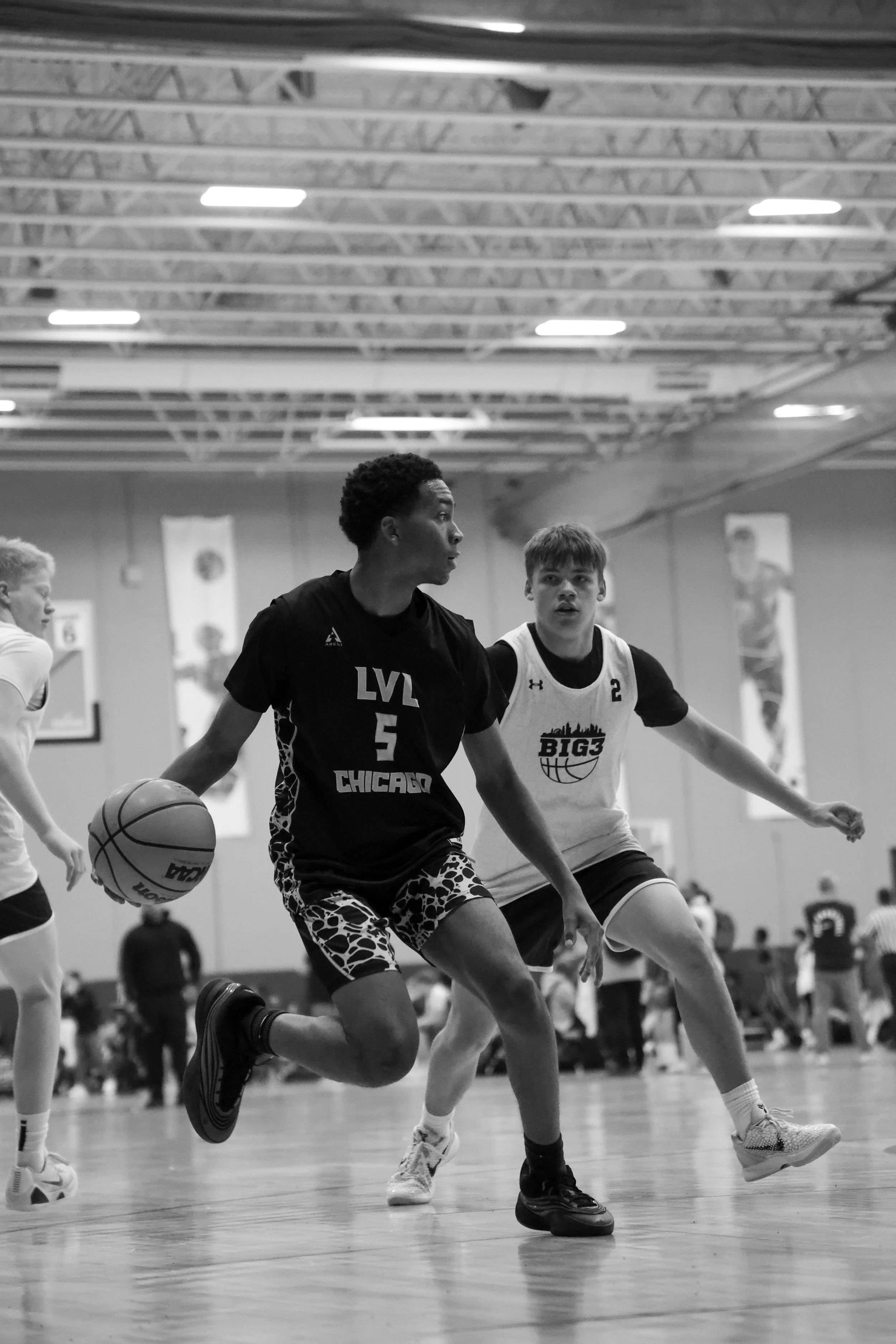 Black and white photo of young athletes playing basketball on an indoor court, with one player dribbling the ball while others are defending.