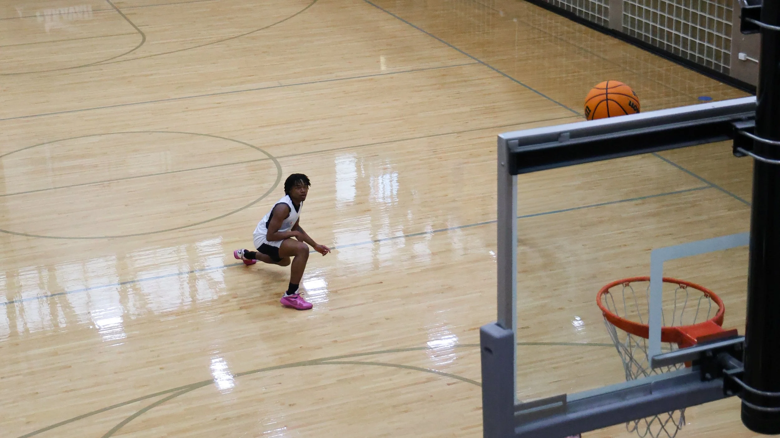 A young basketball player crouching on an indoor basketball court with a basketball in the background and a basketball hoop nearby.