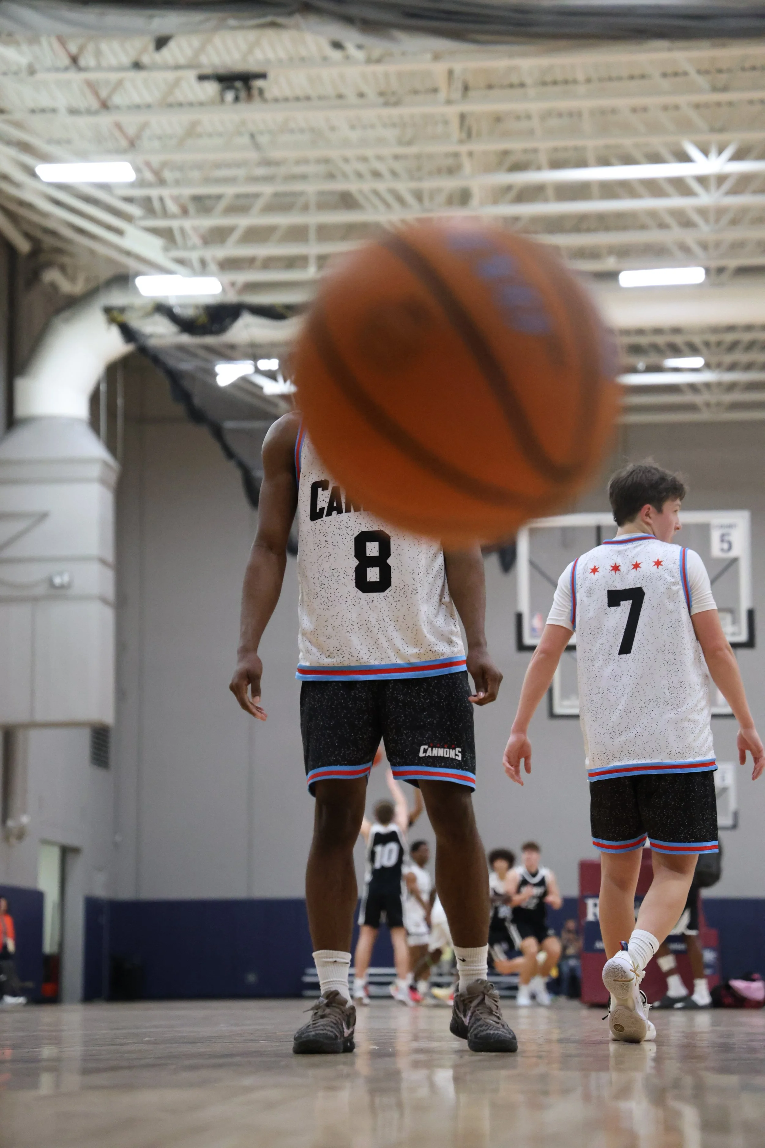 Close-up of two basketball players standing on a gym floor, with the ball in front of the camera. Player on the left wears a jersey with the number 8, and player on the right has the number 7. Other players and a basketball hoop are visible in the background.