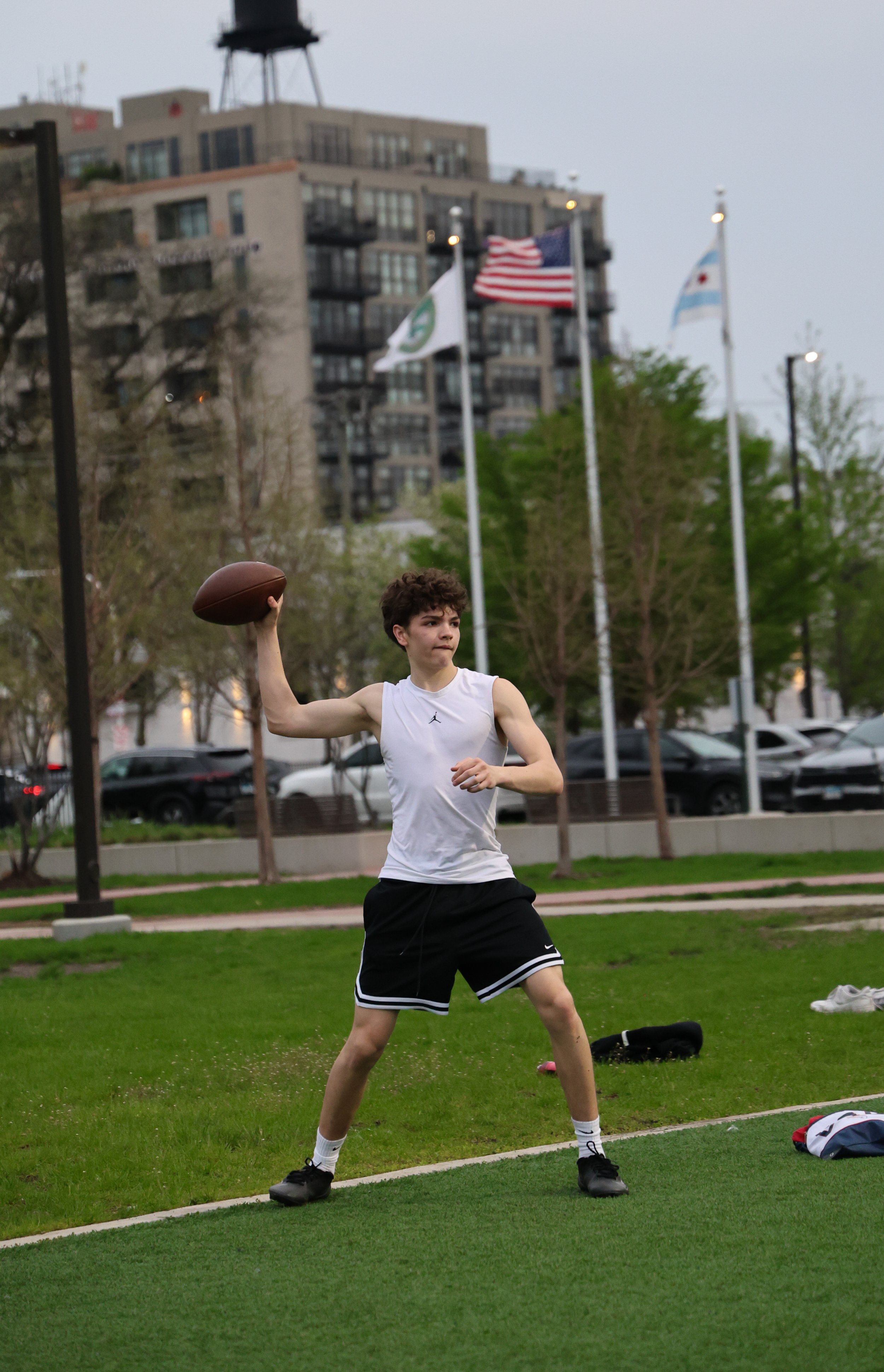 A young male athlete throwing a football in a park with flagpoles, trees, a bench, and a modern building with American flags in the background.