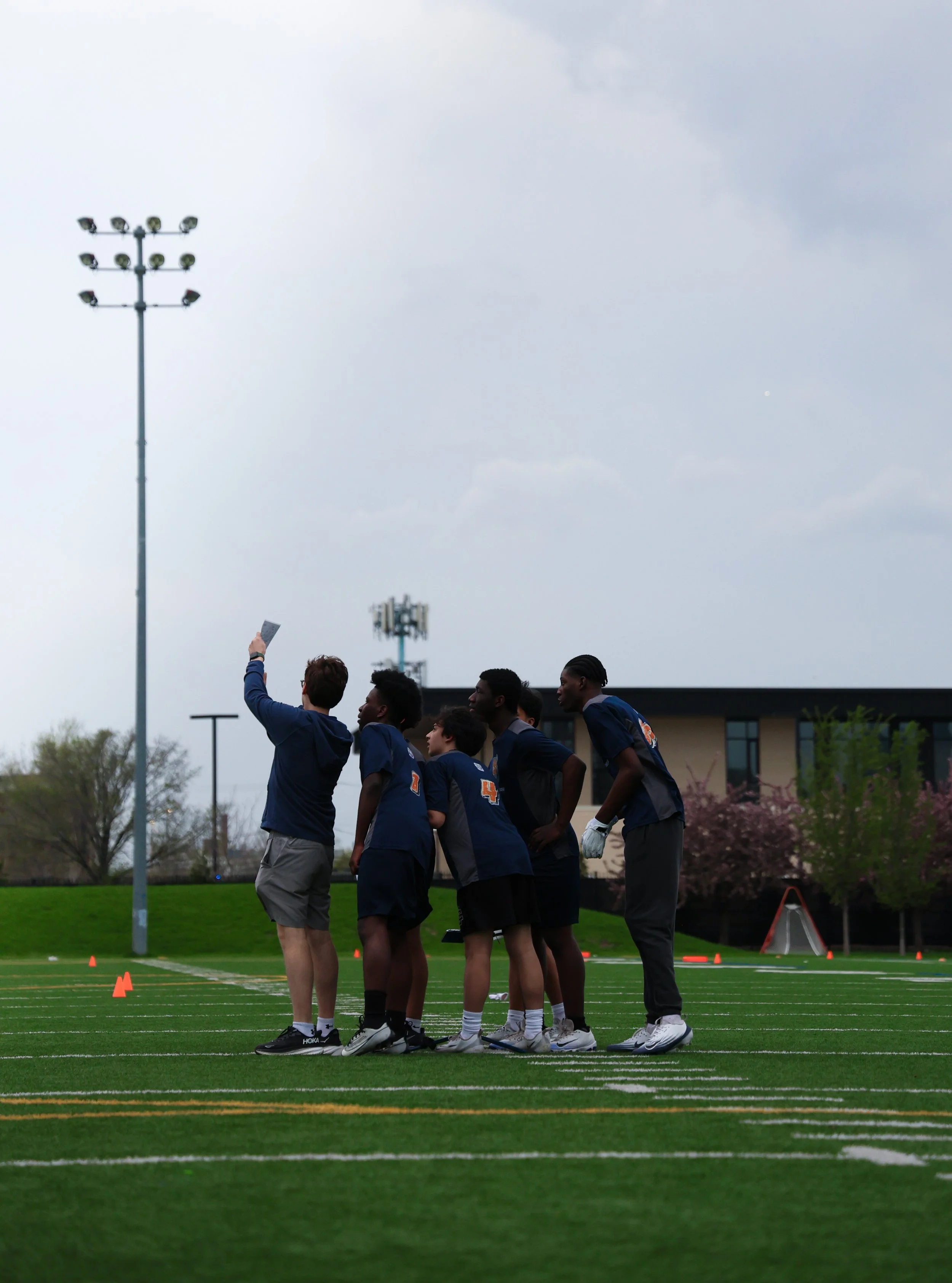 A group of young athletes standing on a football field during a practice or game, with orange cones set up and a cloudy sky above.
