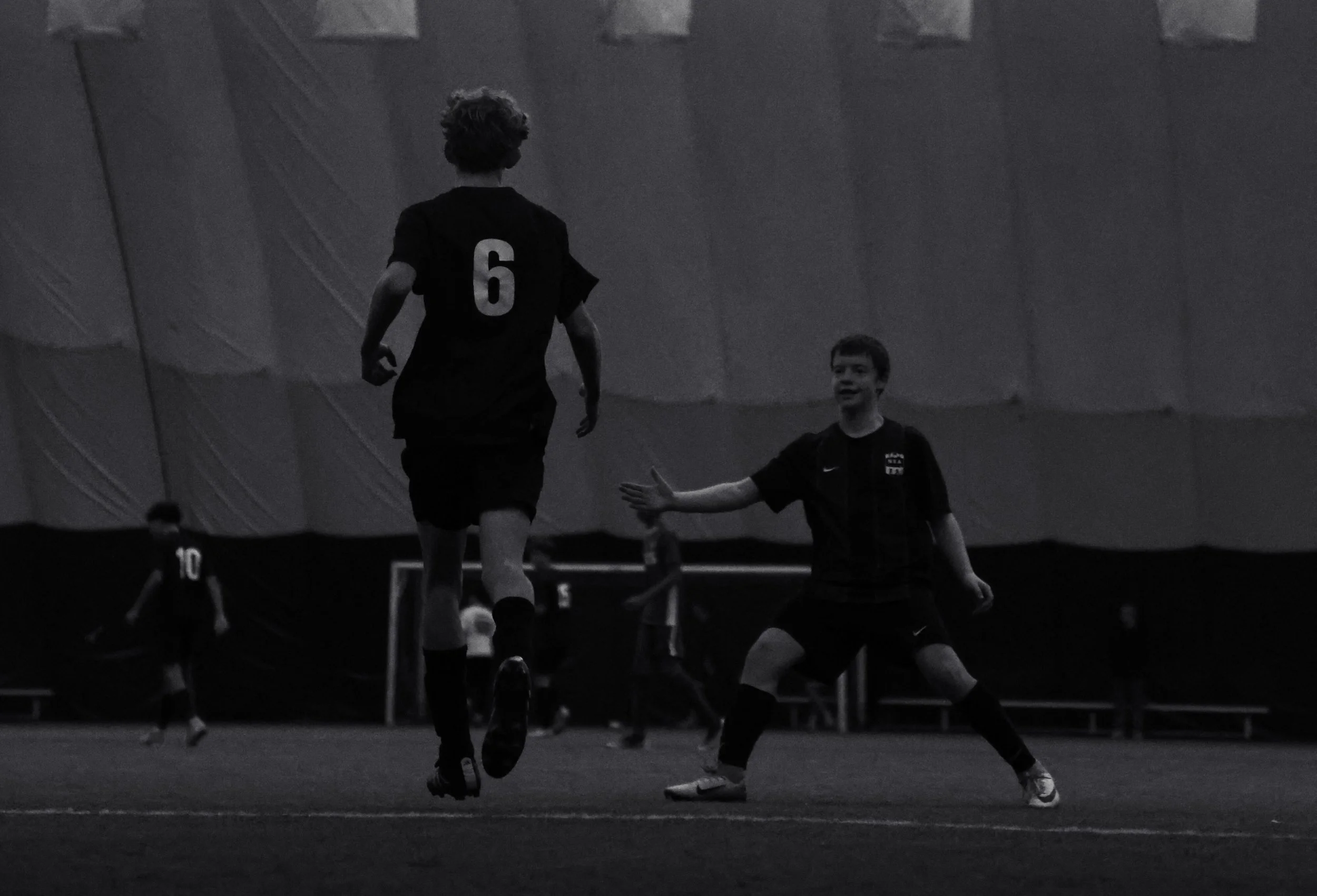 Two young boys playing soccer indoors, with one wearing a black jersey with the number 6 on it. The boy in the jersey is running toward the boy who is extending his hand for a handshake, suggesting a friendly interaction or celebration.