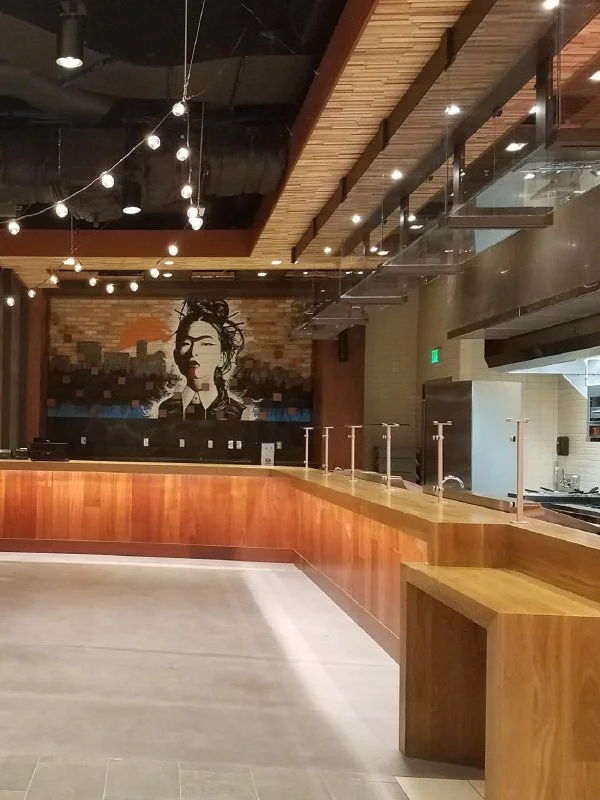 Empty restaurant or cafe interior with wood counter, hanging light bulbs, and artwork of a woman's face on brick wall.