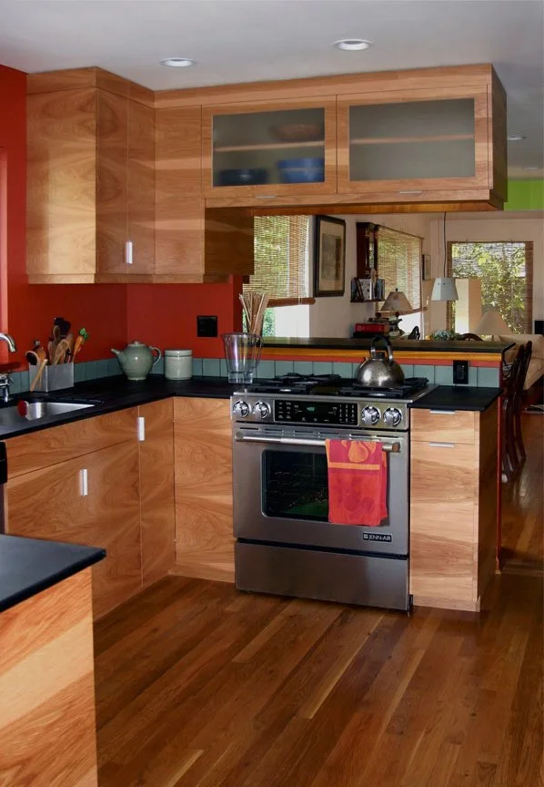 Kitchen with wood cabinets, black countertops, a stainless steel oven with a tea kettle and a towel hanging, and a mirror reflecting a dining area with a window and trees outside.