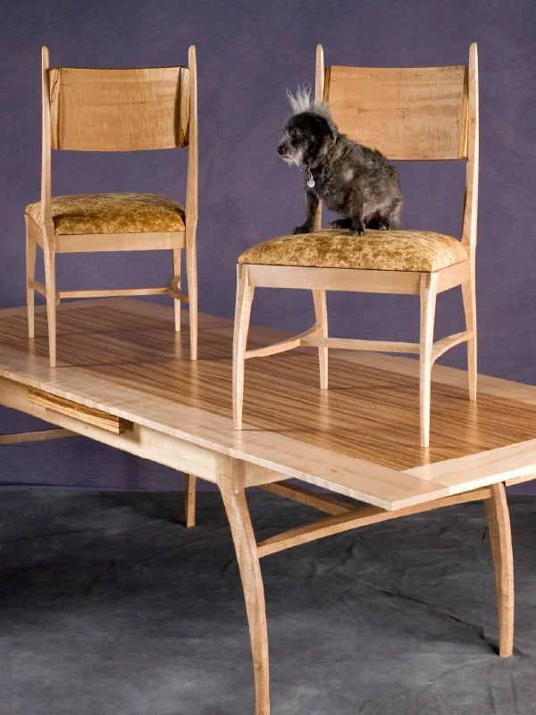 A small dog sitting on a wooden chair with a velvet cushion, positioned on another larger wooden table, against a purple background.