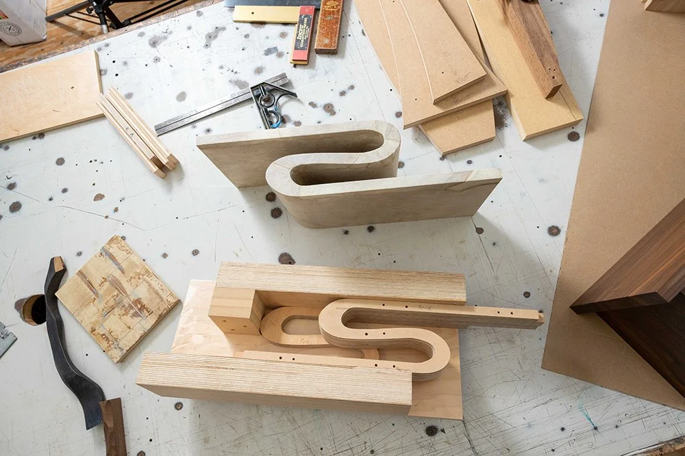 Woodworking workshop with two partially assembled wooden letters 'S' and 'U', surrounded by various wood pieces, tools, and scraps on a worktable.
