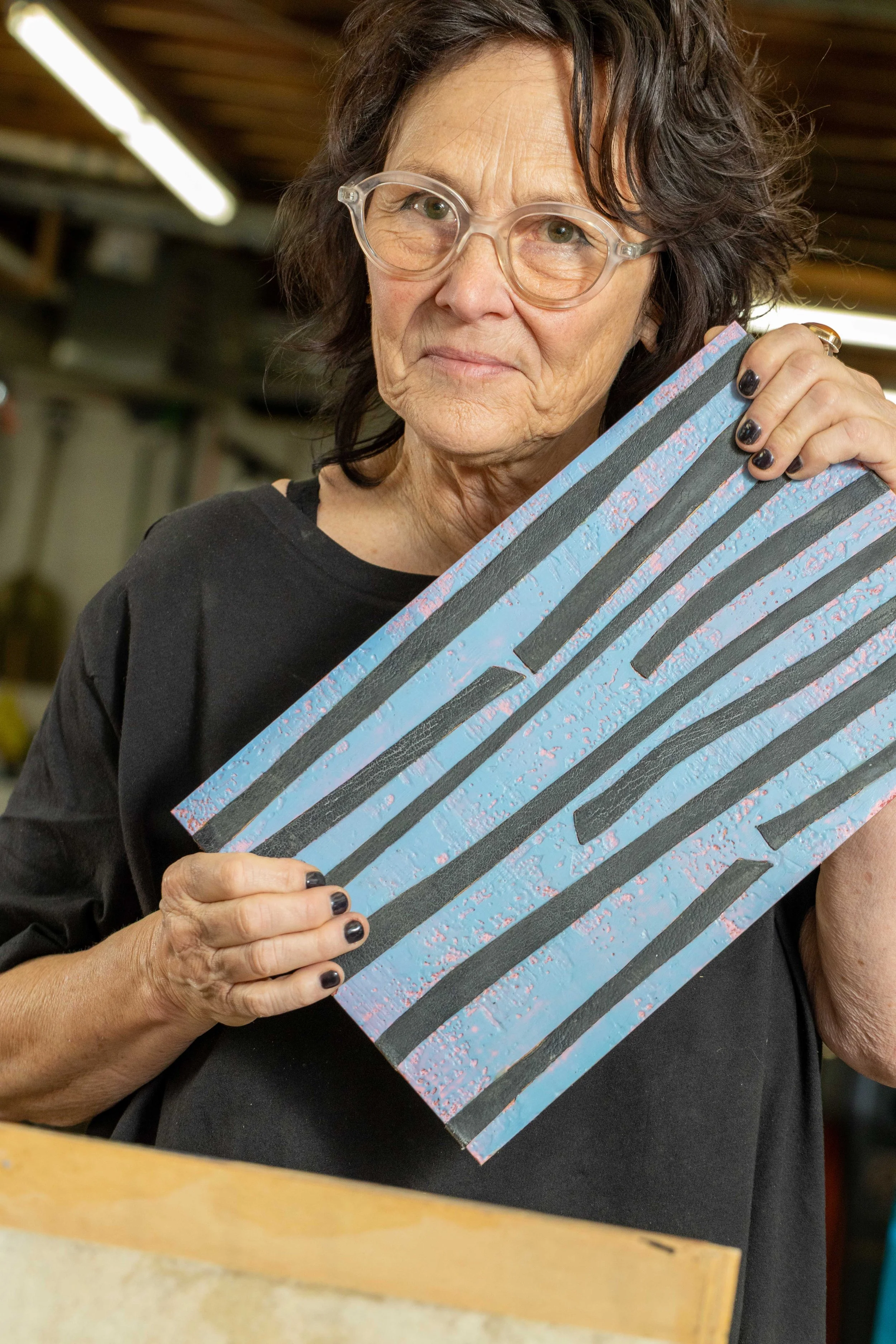 Collette wearing glasses and a black shirt, holding a painted wooden sign with black and pink stripes.
