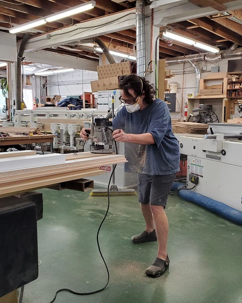 Collette working with woodworking machinery in a woodworking shop, wearing glasses and a mask.