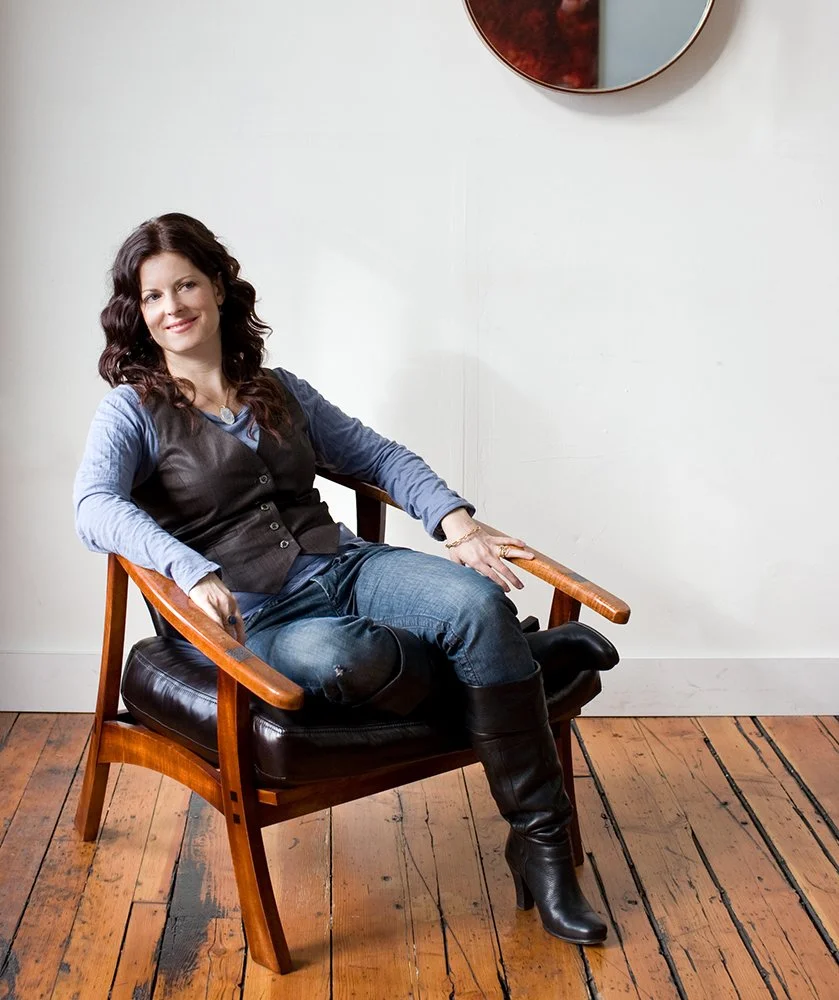 A woman with wavy dark hair sitting on a wooden chair with black leather cushions, wearing a blue shirt, black vest, jeans, and black knee-high boots, in a room with wooden floors and a white wall.