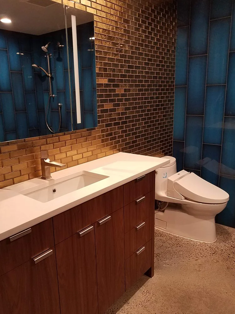 Modern bathroom with wooden vanity, white sink, mirror, shower with blue tiles, brown brick wall, and a high-tech toilet.