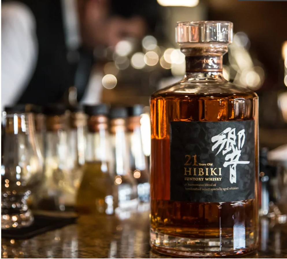 A bottle of Hibiki 21 Year Old Japanese whisky sits on a polished bar, with glassware and bottles softly blurred in the background.