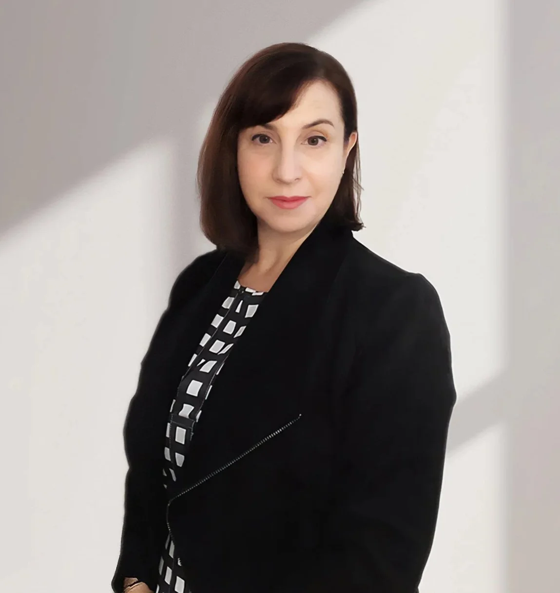A woman with shoulder-length brown hair wearing a black blazer and a black-and-white checkered blouse, standing against a white wall with a shadow.