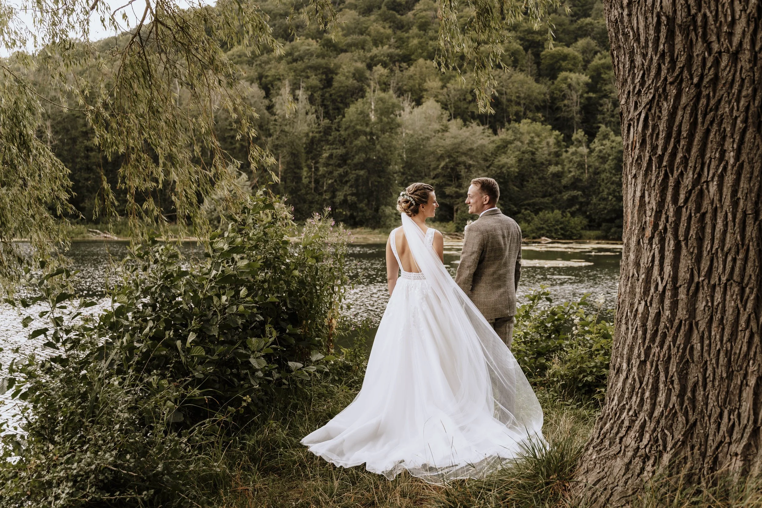 Ein Brautpaar steht am Ufer eines Sees in einem Wald. Die Braut trägt ein langes weißes Brautkleid mit Schleppe, der Bräutigam trägt einen grauen Anzug. Sie schauen sich liebevoll an, umgeben von Bäumen und Natur.