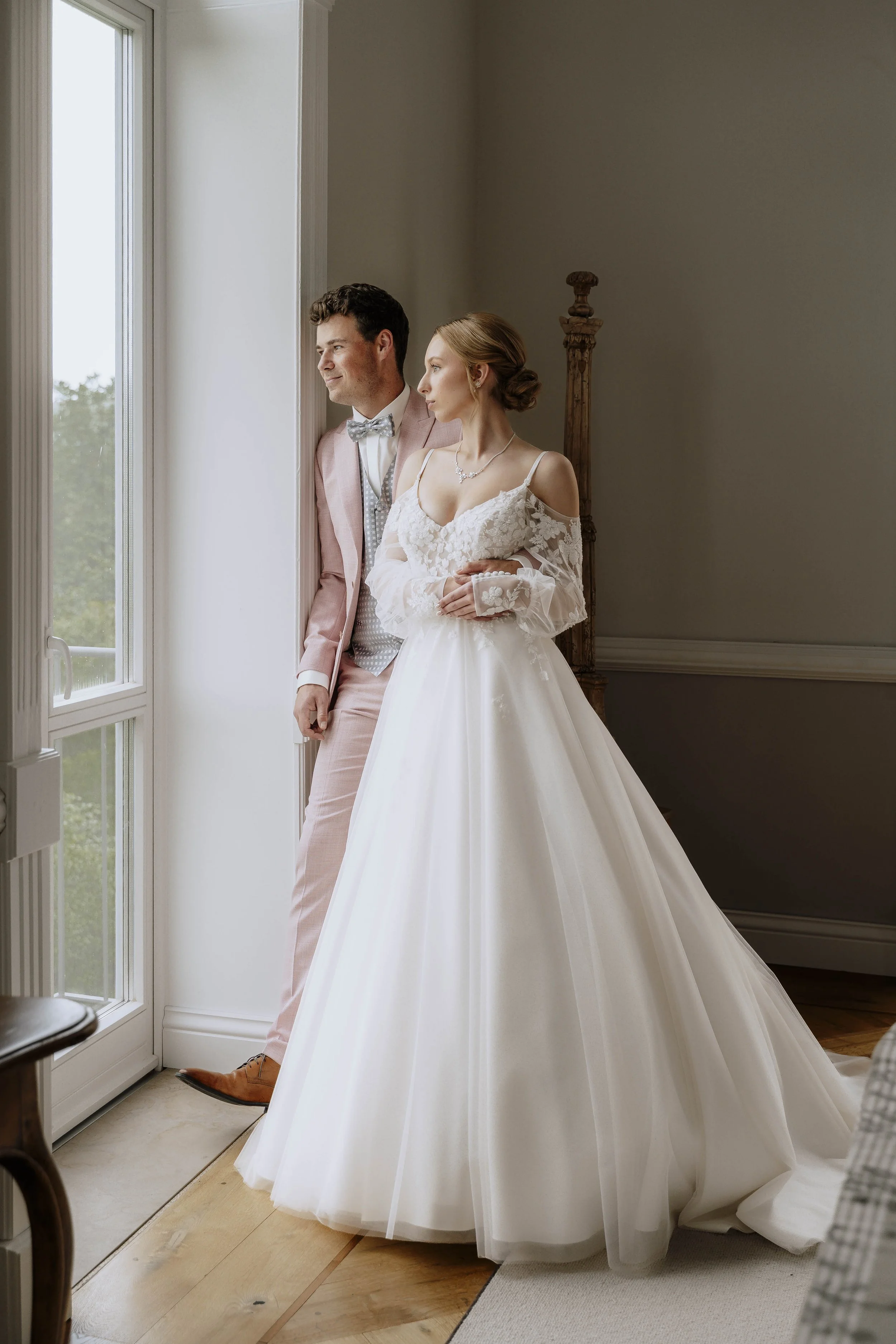 Braut in Hochzeitskleid und Bräutigam in Anzug stehen neben einem großen Fenster und schauen nach draußen.