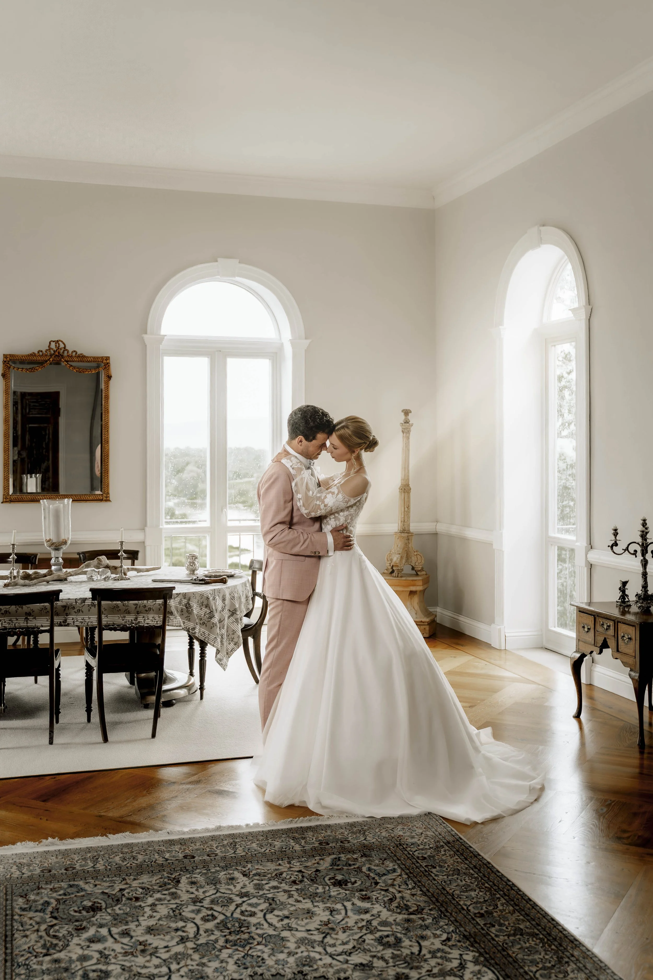 Ein Brautpaar steht in einem hellen, eleganten Raum, umarmt sich und schaut sich liebevoll an. Das Brautkleid ist lang und weiß, das Kleid des Bräutigams ist hellrosa. Der Raum hat große Fenster, edle Möbel und Dekorationen im klassischen Stil.