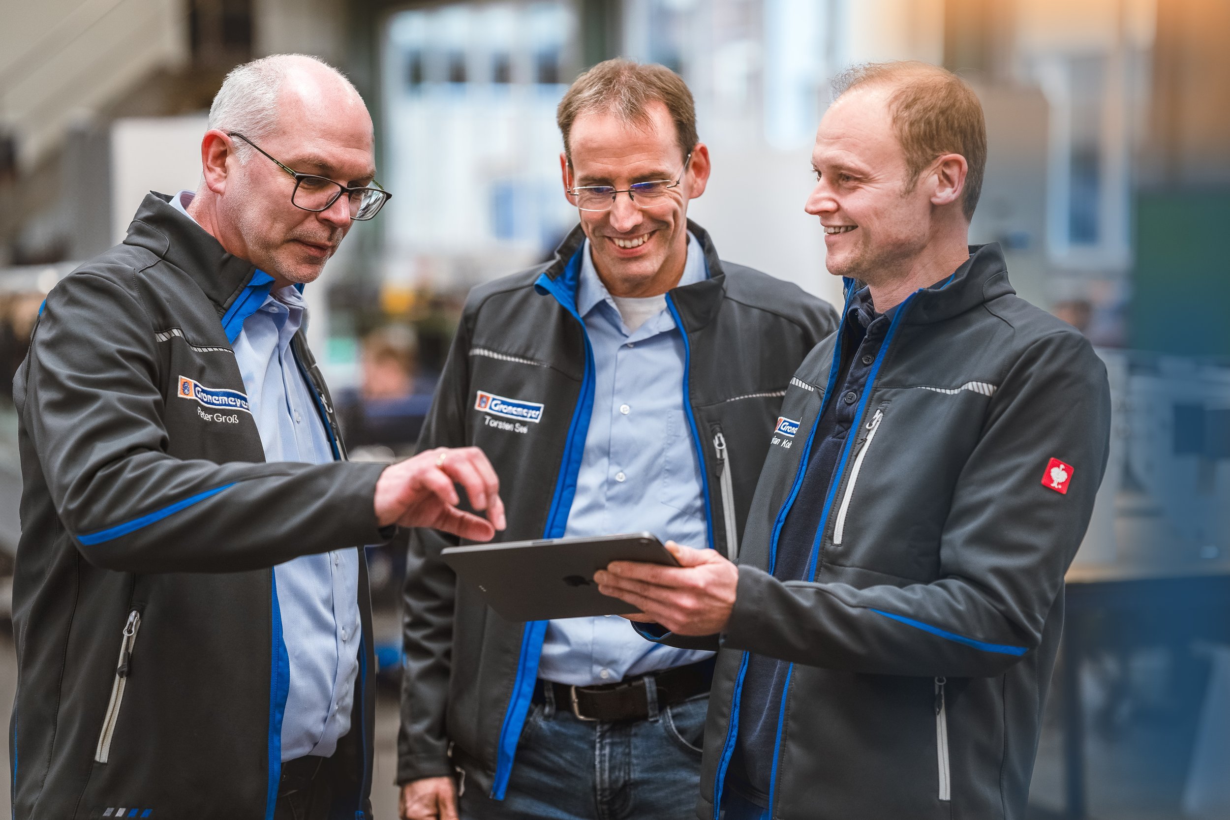 Drei Führungskräfte der Gronemeyer Maschinenfabrik diskutieren Daten auf einem Tablet in der Produktionshalle – Corporate Teamfoto für Imagekommunikation und Website