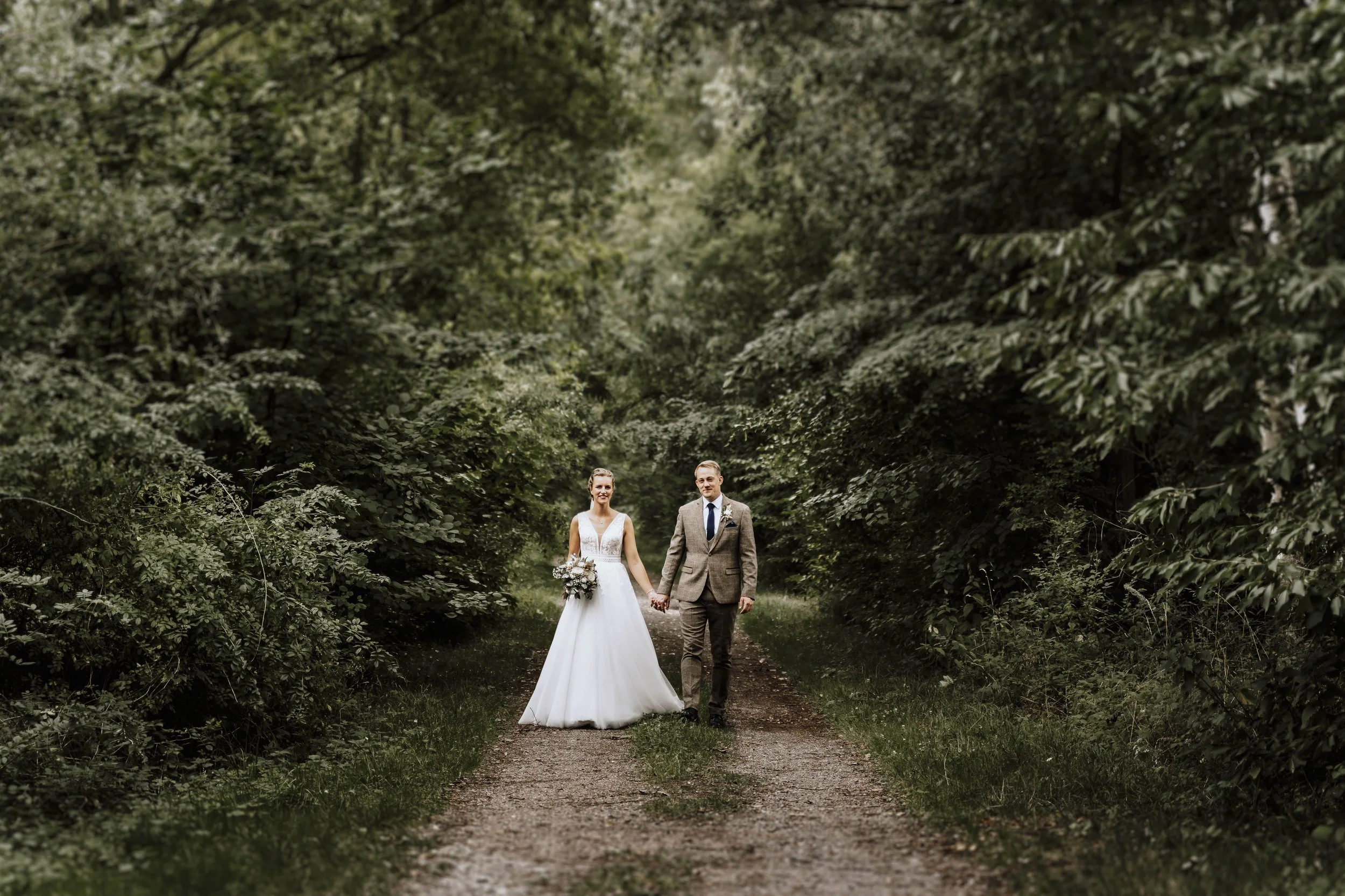 Ein Hochzeitspaar, eine Frau in weißem Hochzeitskleid und ein Mann in Anzug, geht Hand in Hand auf einem Naturweg durch einen grünen Wald.