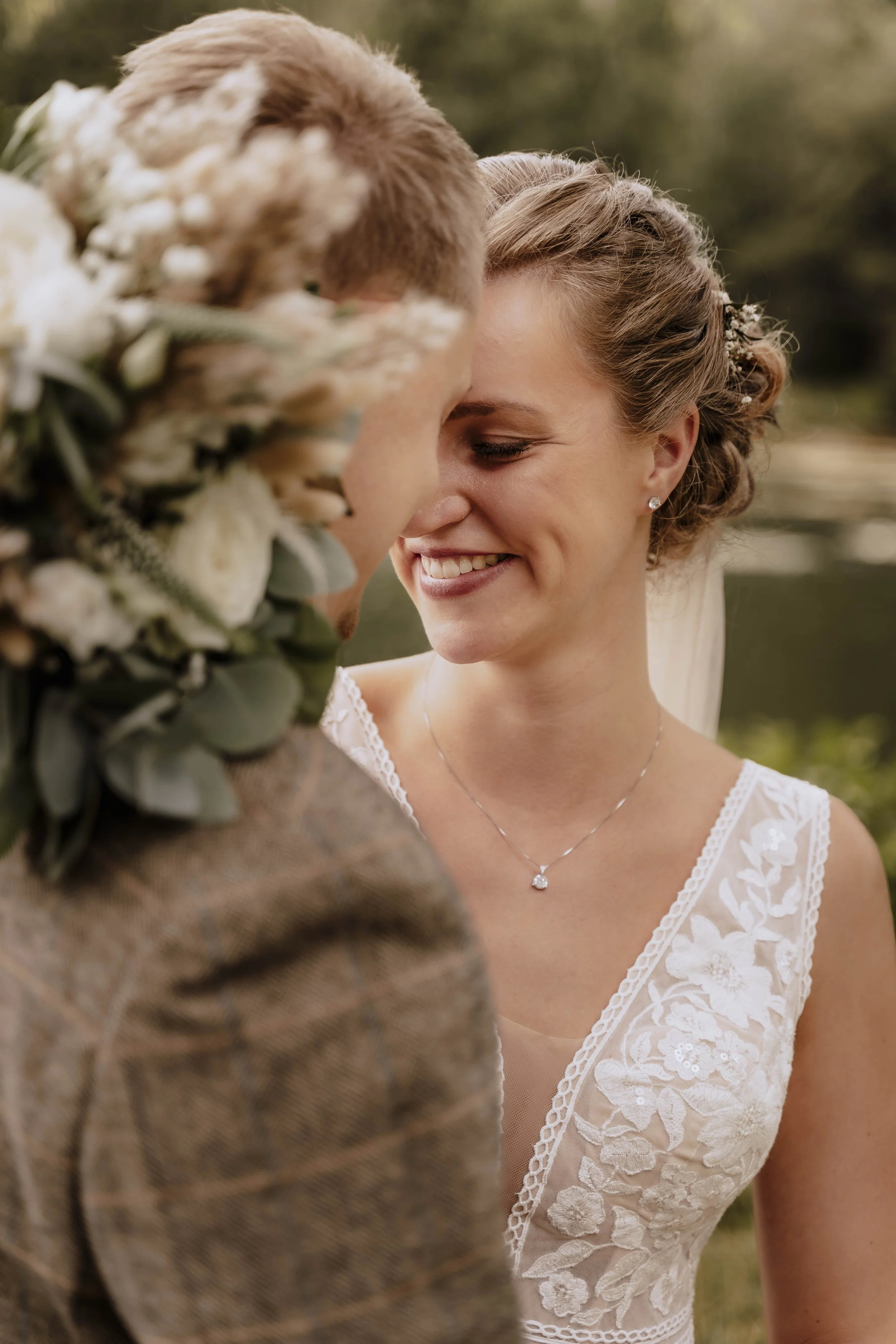 Ein Brautpaar bei einer Hochzeit, Nahaufnahme ihres Gesichts, die Braut lächelt, trägt eine weiße Spitze Brautkleid und Schmuck, im Hintergrund unscharfer Naturbereich.