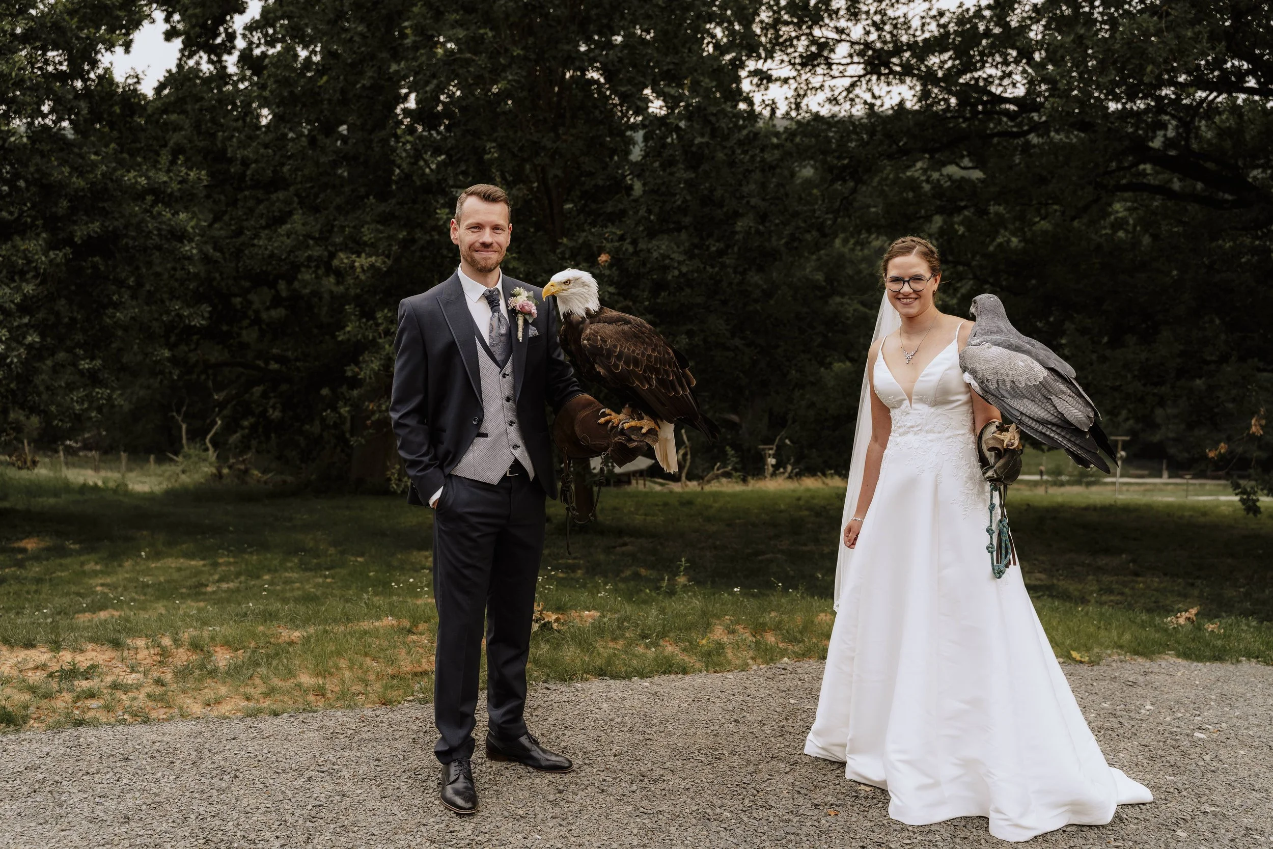 Brautpaar posiert gemeinsam mit zwei Greifvögeln (Weißkopfseeadler und Bussard) auf dem Kiesweg – einzigartige Falknerei-Hochzeitsfotos