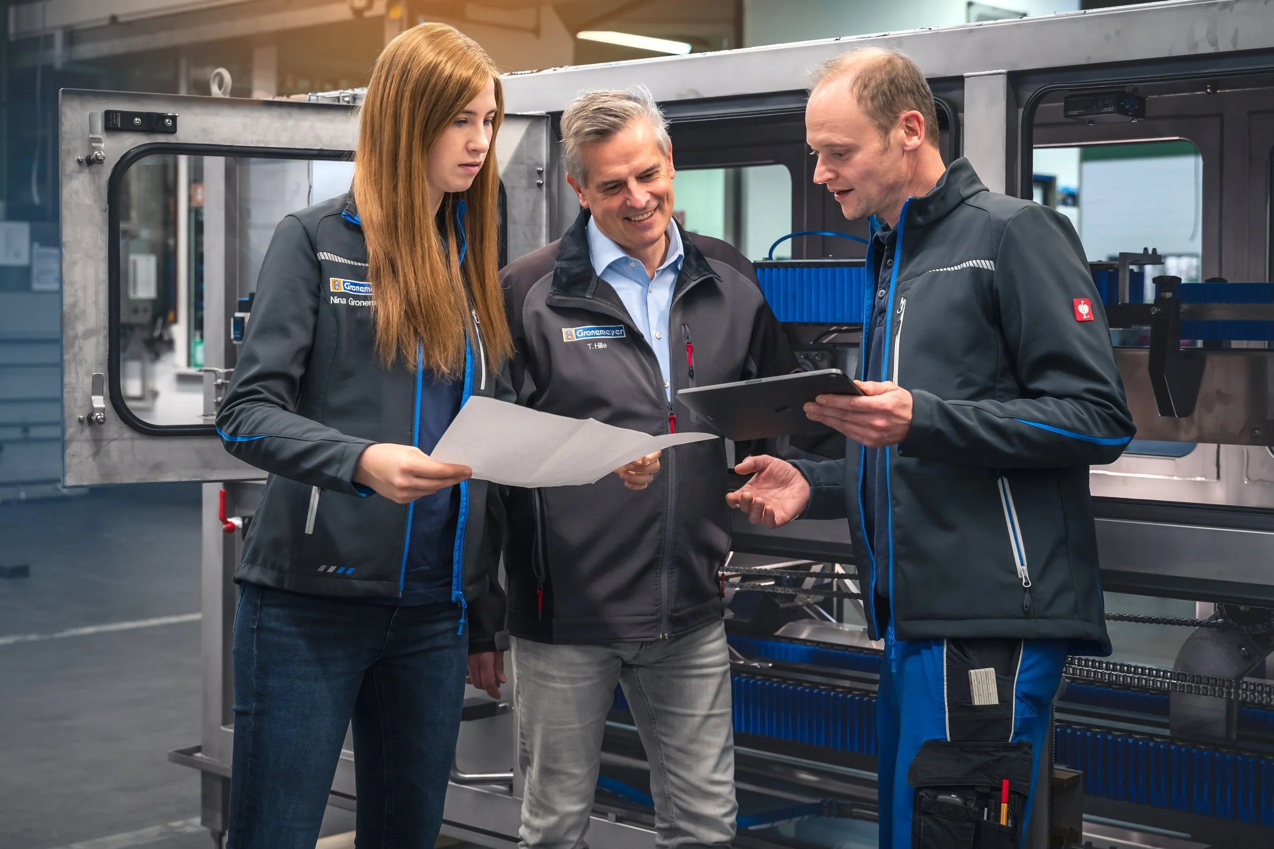 Corporate Reportage Maschinenfabrik Gronemeyer: Drei Mitarbeitende besprechen Unterlagen und Tablet vor industrieller Fertigungsanlage – Unternehmensreportage Kreis Höxter