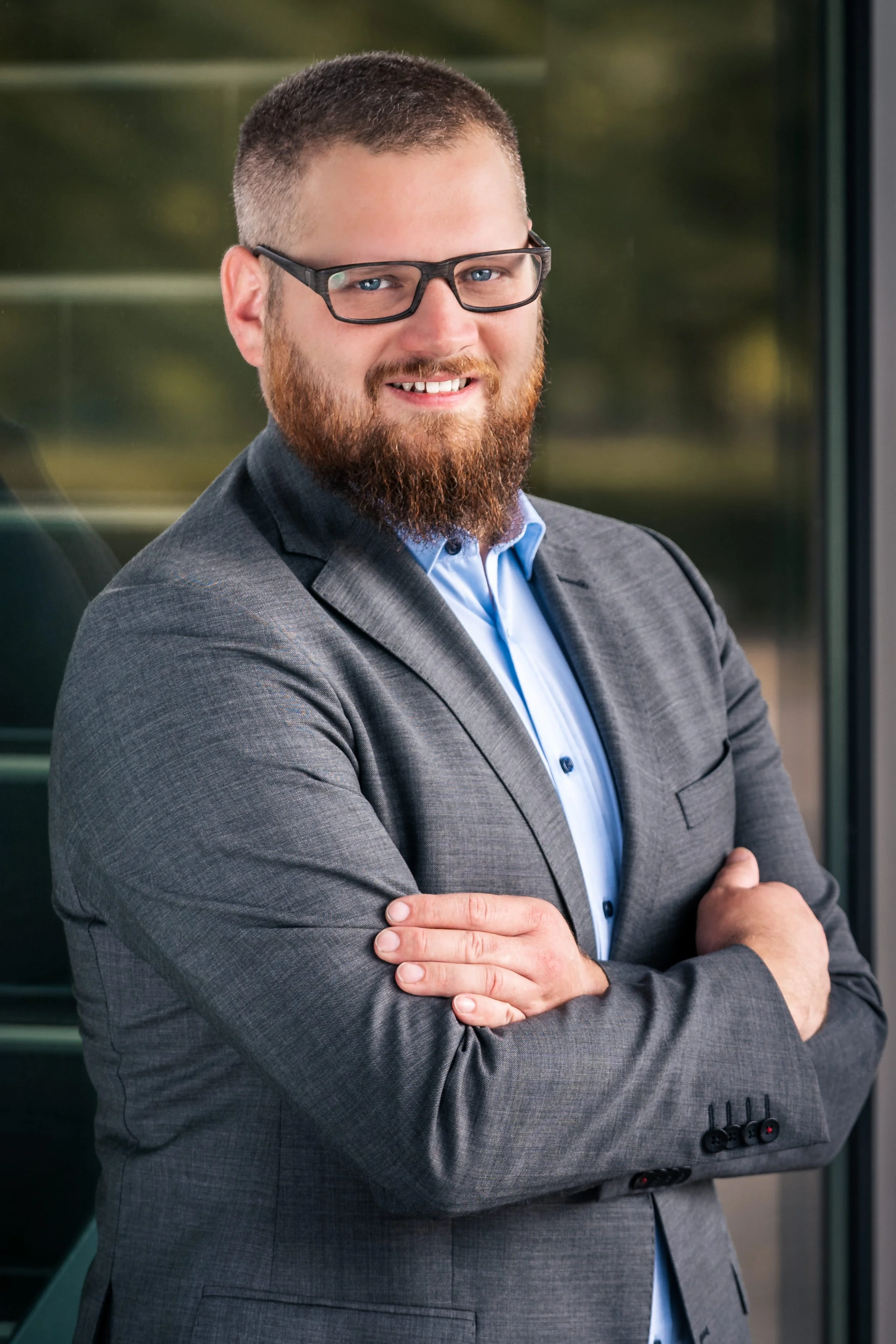 Business-Headshot eines lächelnden Managers mit Bart und Brille, anlehend an Glasfassade im Außenbereich – professionelle Businessfotografie Niklas Schulz Photography