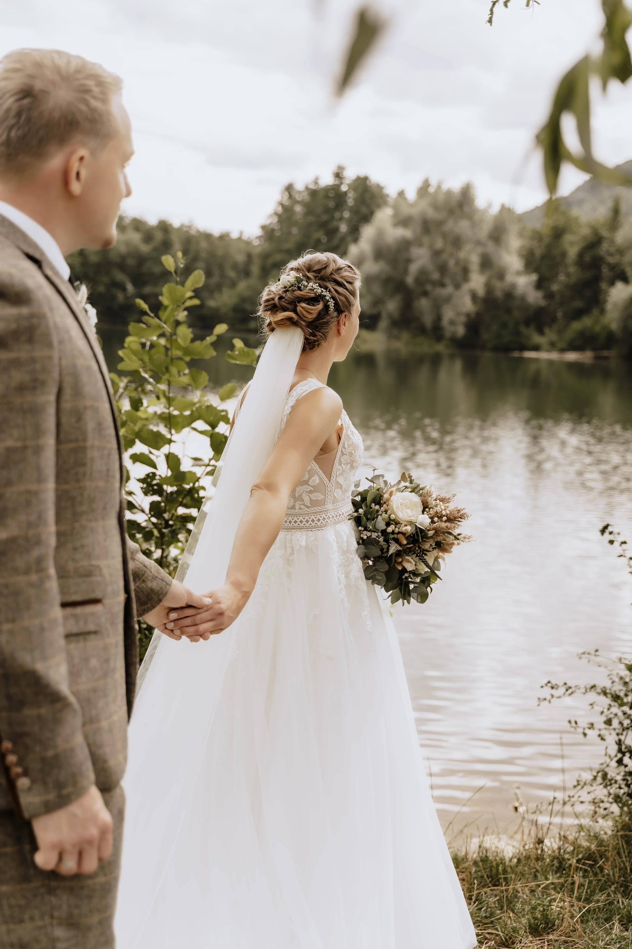 Ein Brautpaar hält Händchen an einem Flussufer, die Braut trägt ein weißes Kleid mit Spitze und einen Brautstrauß, während der Bräutigam einen braunen Anzug trägt, in einer ruhigen, naturbelassenen Umgebung mit Bäumen und Wasser im Hintergrund.