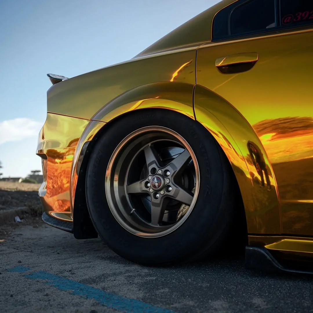 Close-up of a gold-colored sports car's front left wheel and body, reflecting sunset sky and nearby landscape.