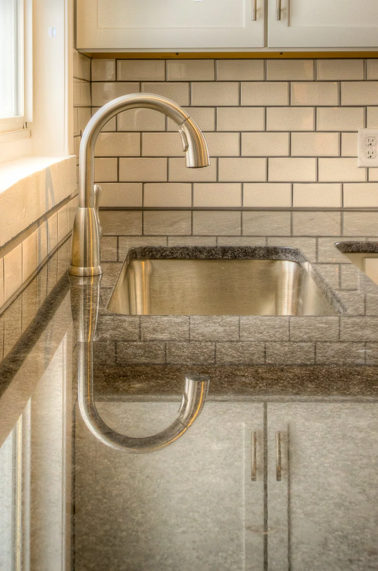 Kitchen countertop with a stainless steel sink and a modern chrome faucet, reflected on the black granite surface. Light-colored tile backsplash and wall with an electrical outlet.