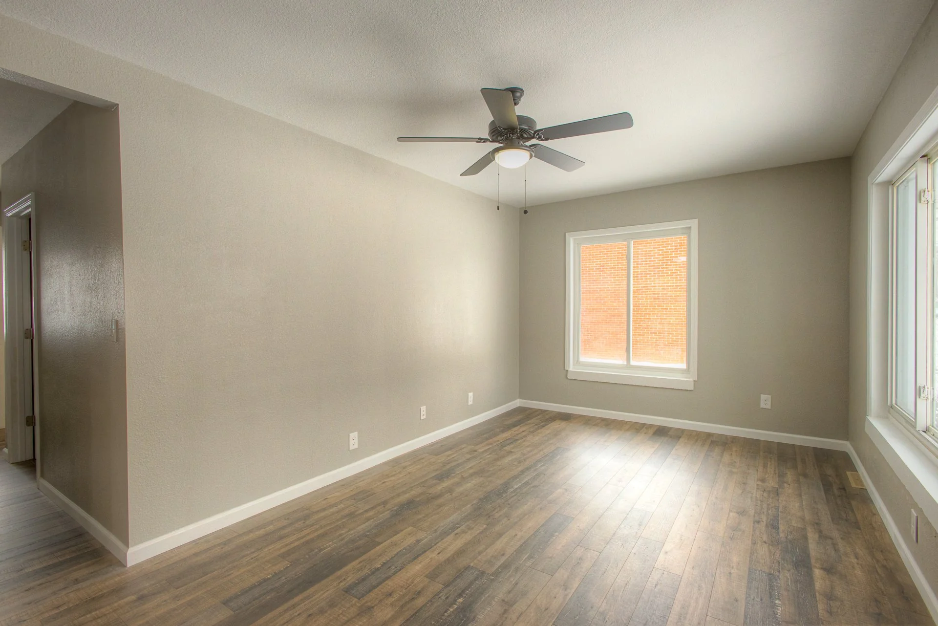 Empty living room with wood flooring, beige walls, a ceiling fan, a window, and a sliding glass door.