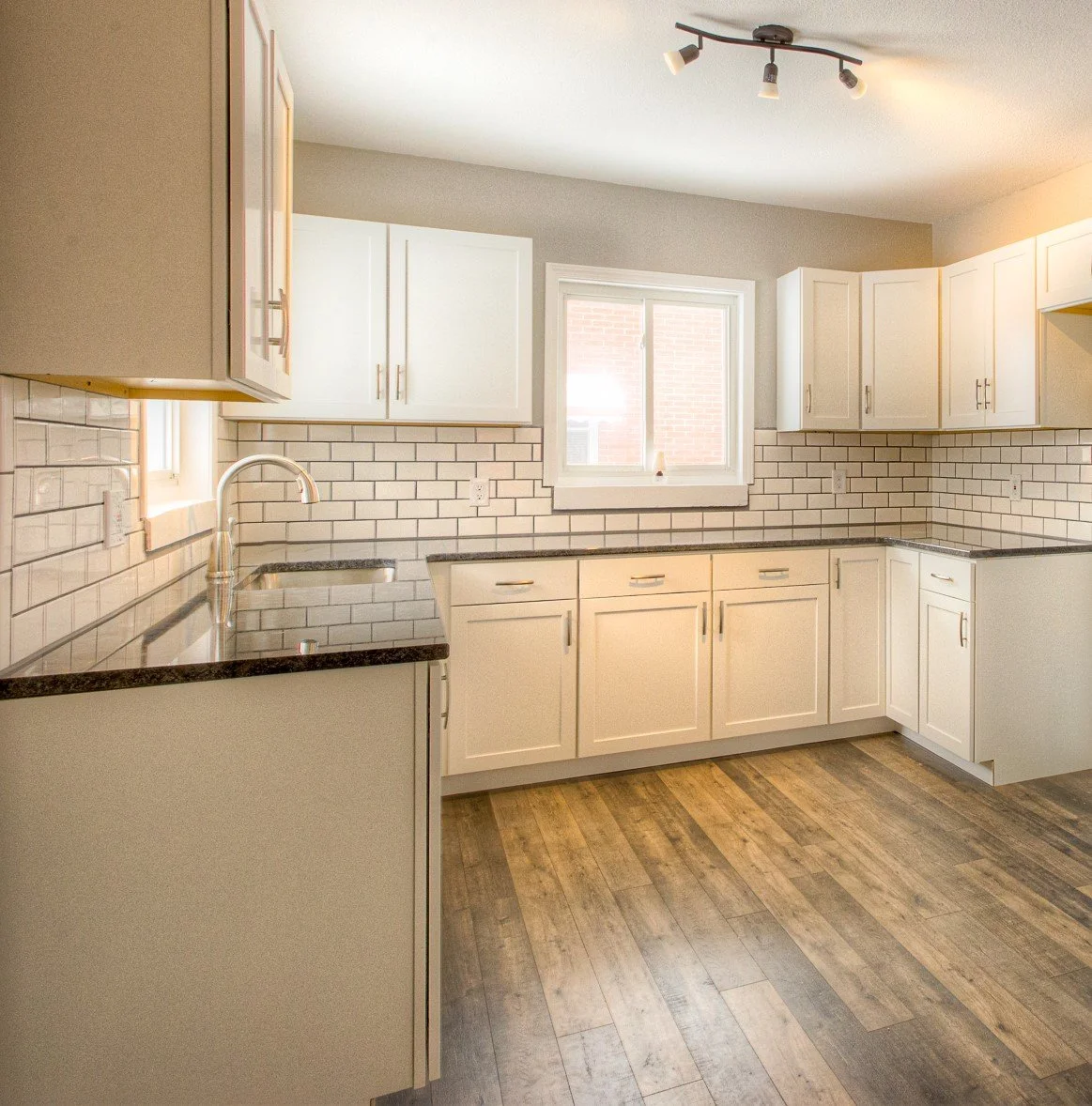 Kitchen with white cabinets, black countertop, white subway tile backsplash, window above the sink, and wooden flooring.