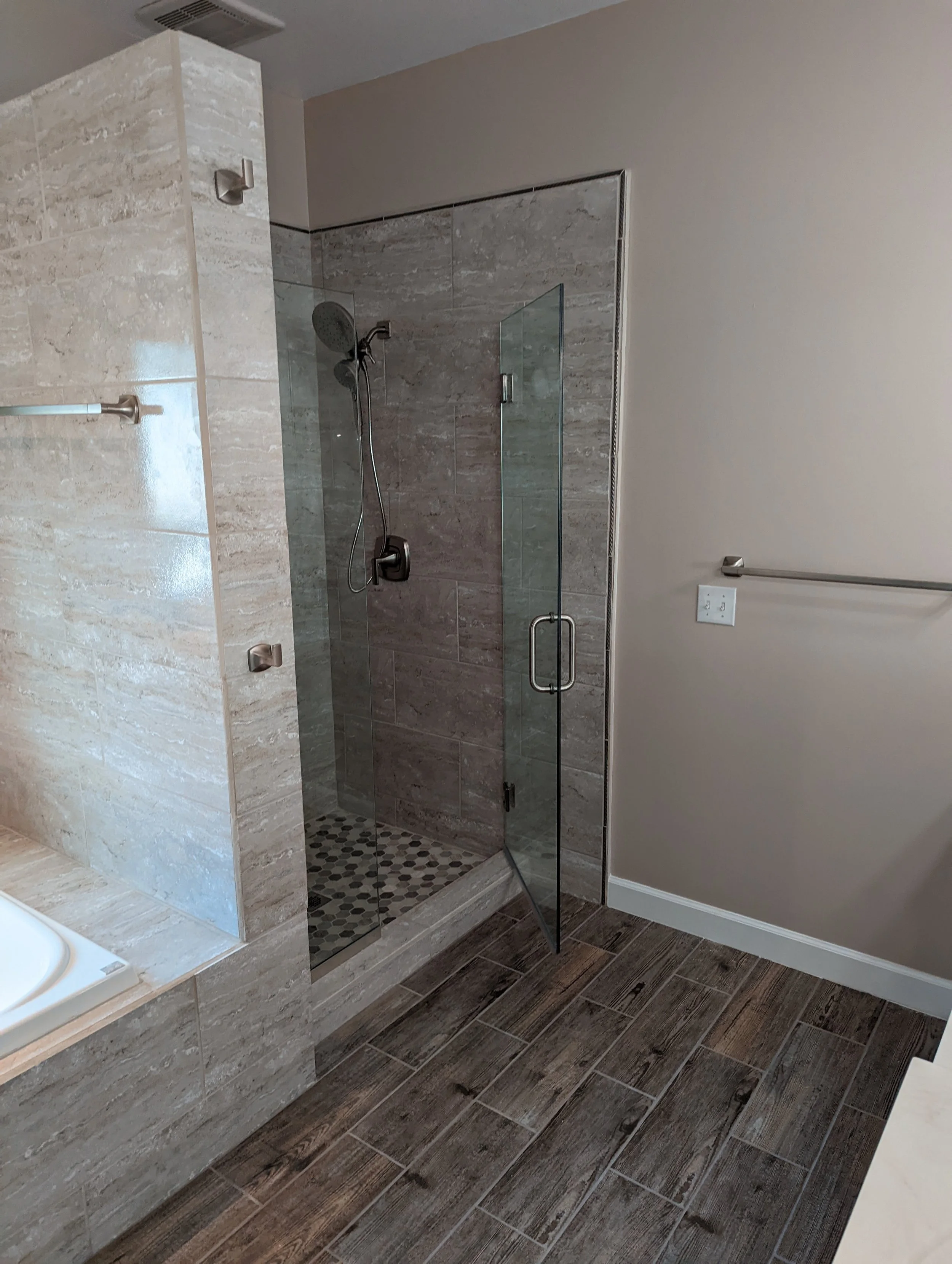 A modern bathroom with a glass-enclosed shower, beige and gray tiled walls, dark wood-look tile flooring, and a towel bar on the right wall.