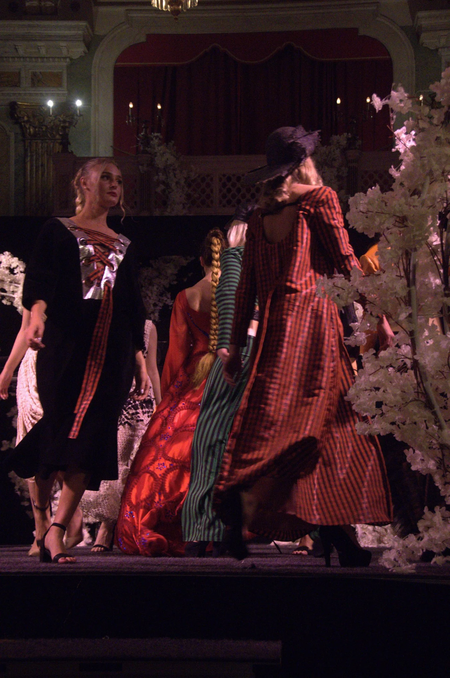Women dressed in vintage-style clothing on a stage, surrounded by white flowers, with dark red curtains and ornate architecture in the background.