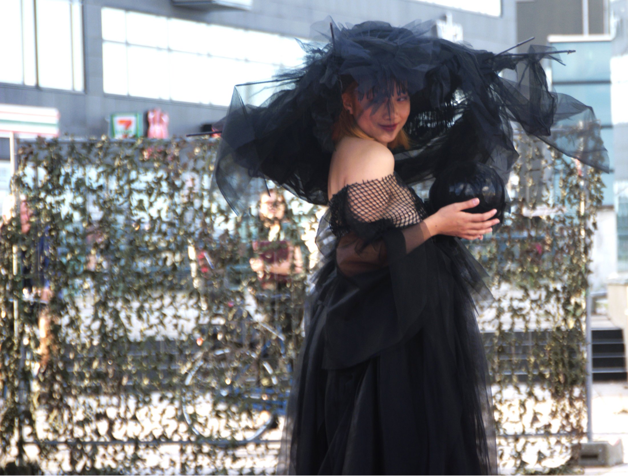 A person in a black tulle dress and fishnet sleeves holding a black heart-shaped object outdoors, with a hedge and building in the background.