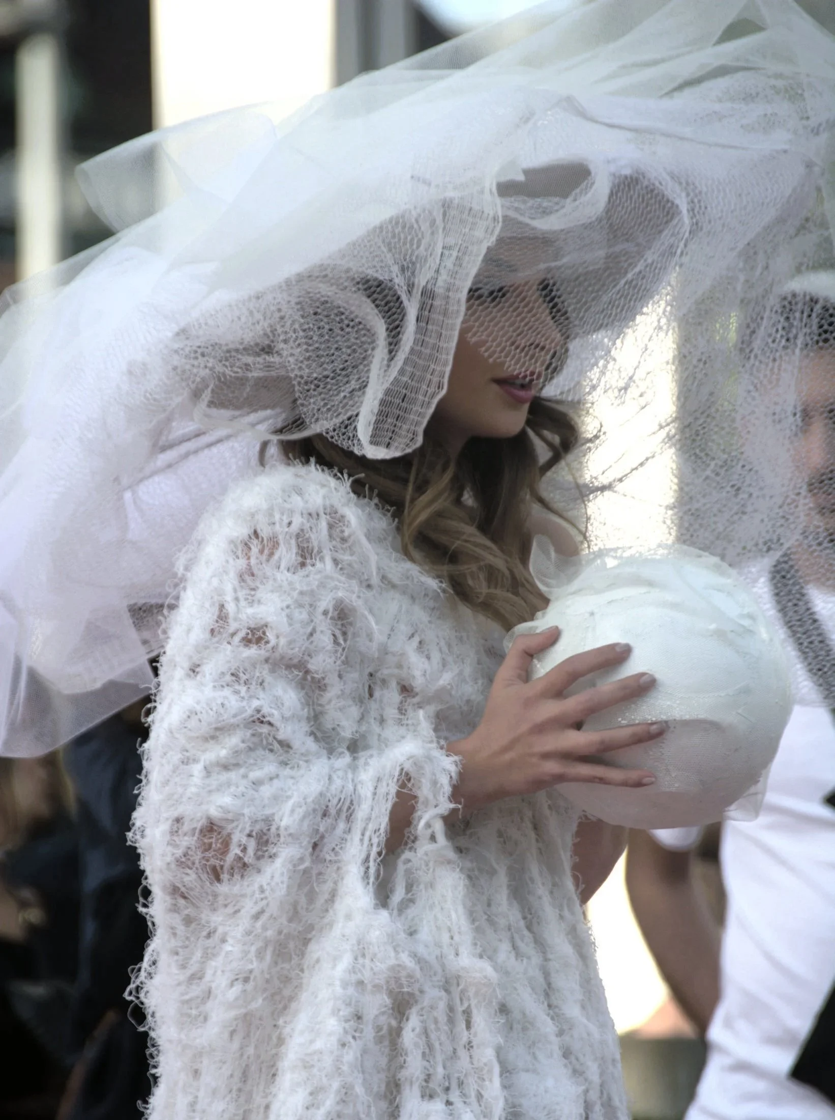 A woman wearing a white, lace dress and a wide-brimmed veil hat holds a white bouquet, surrounded by people at an outdoor event.