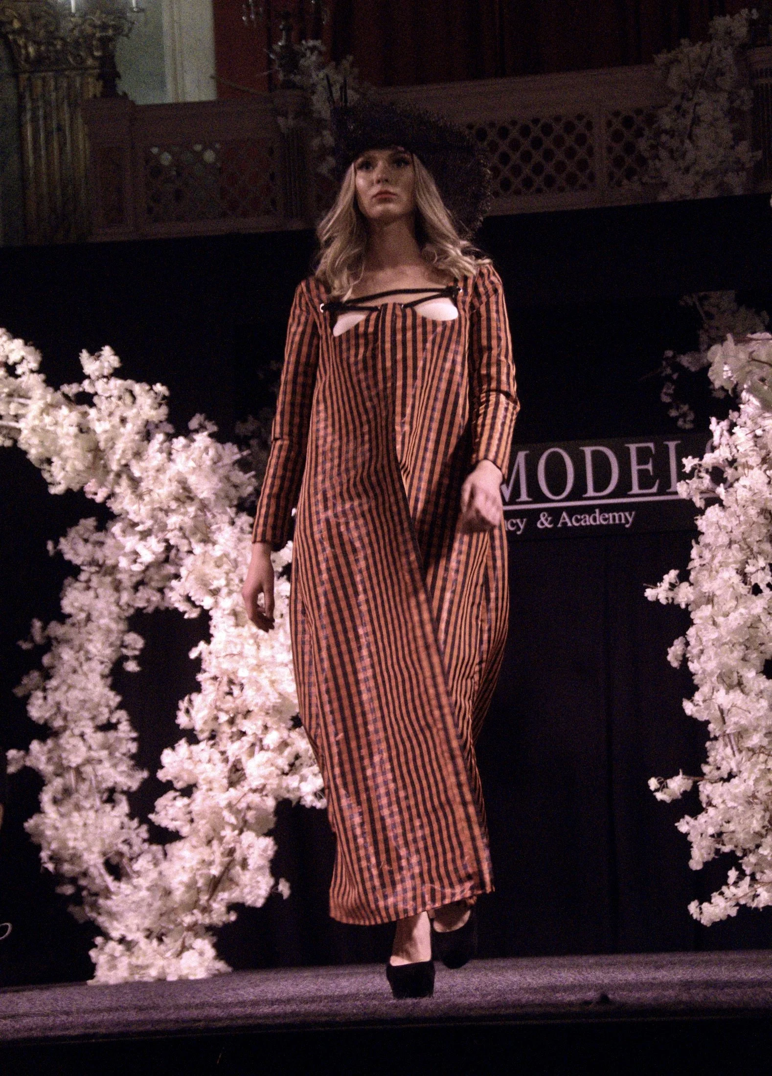 A woman walking on stage during a fashion show, wearing a long, striped dress with cut-out details on the chest, a large black floppy hat, and black heels. The stage is decorated with white flowers and there is a black backdrop with the partially vis