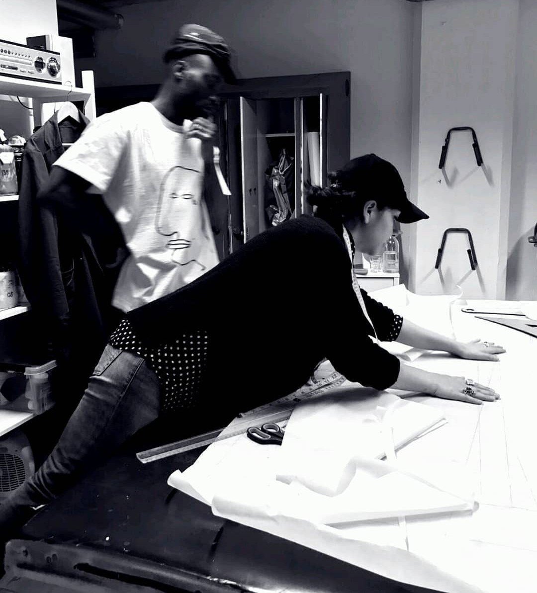 Two people working on a large sheet of paper or fabric on a table in a room, with shelves and tools in the background.