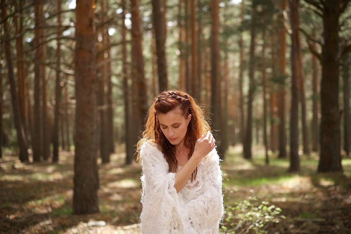 A woman with long, wavy brown hair and a white, fluffy sweater standing in a sunlit forest with tall trees.
