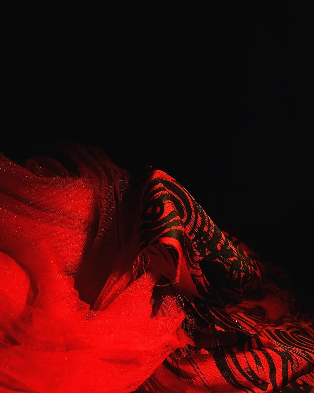 Close-up of red and black patterned fabric illuminated by red light, with a sheer red material in the foreground.
