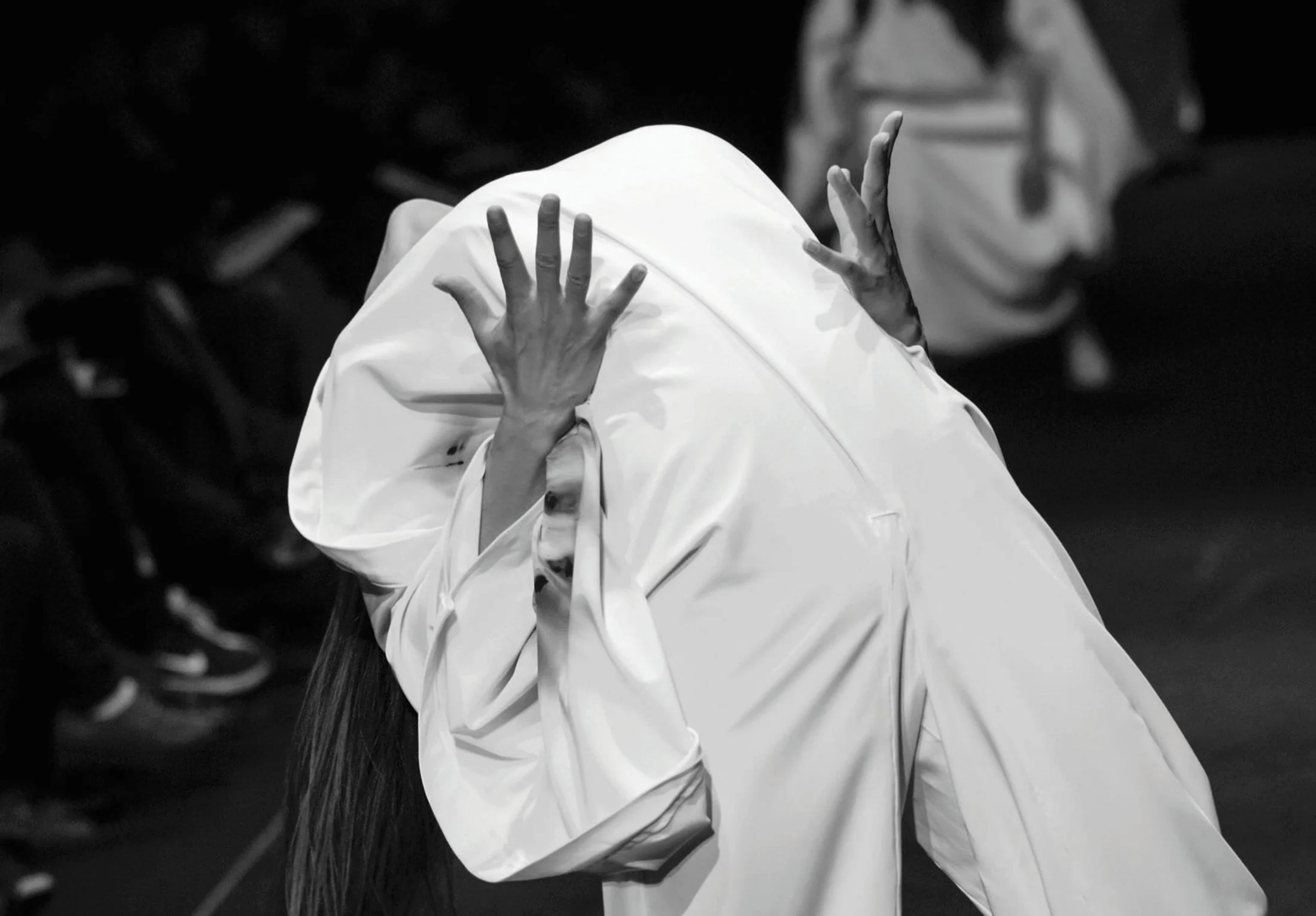 Black and white photo of a person with long hair, wearing a white garment, raising their hand with fingers spread.