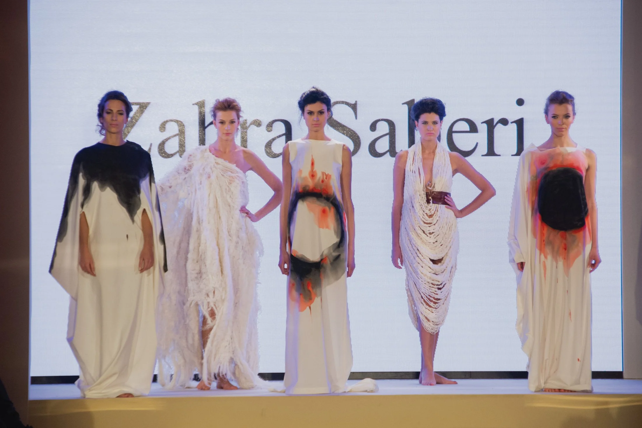 Five women walk down a runway, showcasing designer dresses at a fashion show, with a large white screen behind them displaying the words 'ZahraSaher' in glittery letters.