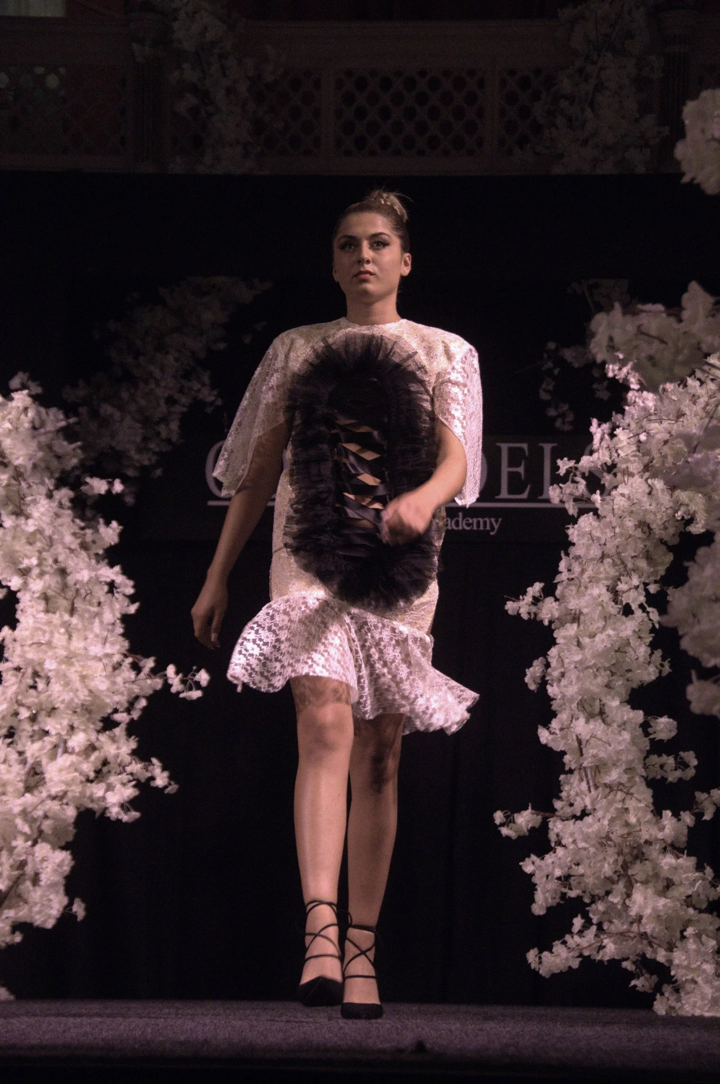 A female model walking on a runway surrounded by pink blossoms, wearing a lace dress, black lace-up high heels, and holding a black feathered fan.