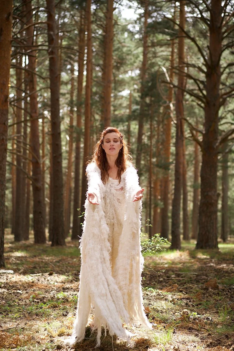 A woman in a long, white, fuzzy dress stands with outstretched arms in a sunlit forest surrounded by tall pine trees.