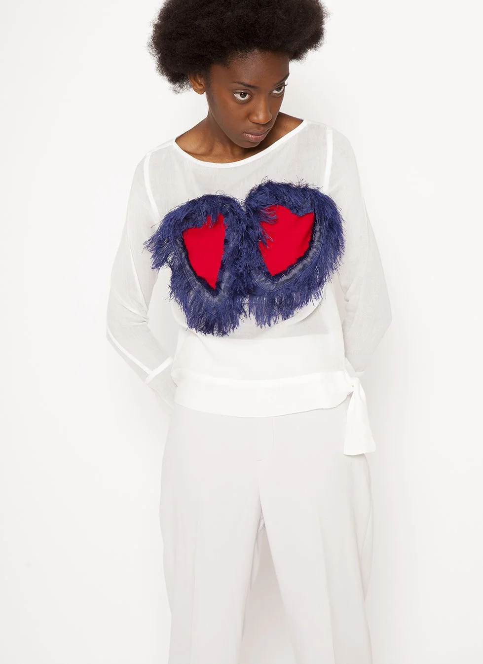 A woman wearing a white outfit with a colorful, feathered heart embellishment on her chest, looking down with a neutral expression, against a plain white background.