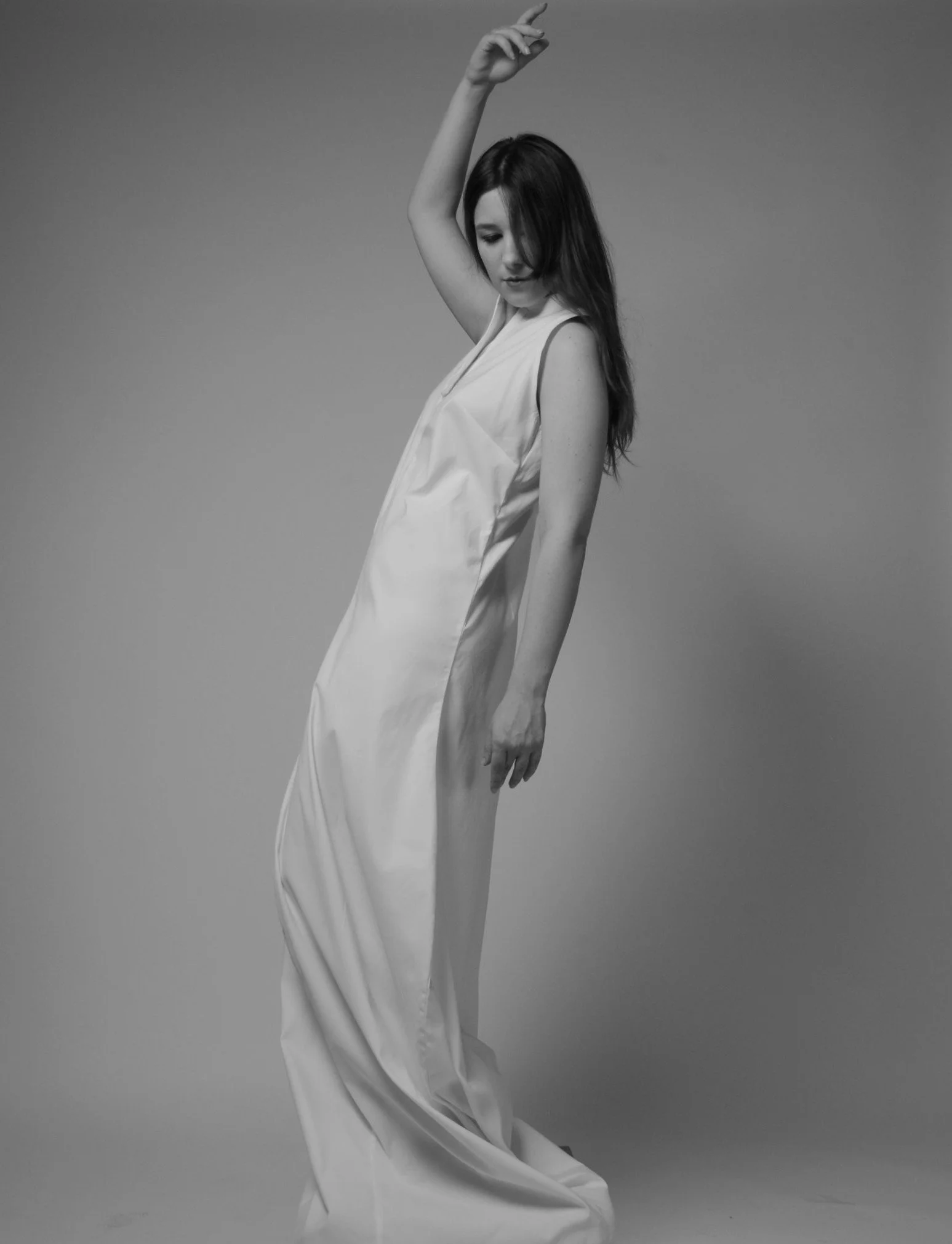 A woman with long dark hair poses in a sleeveless white dress against a plain gray background, with her eyes closed and one arm raised above her head.