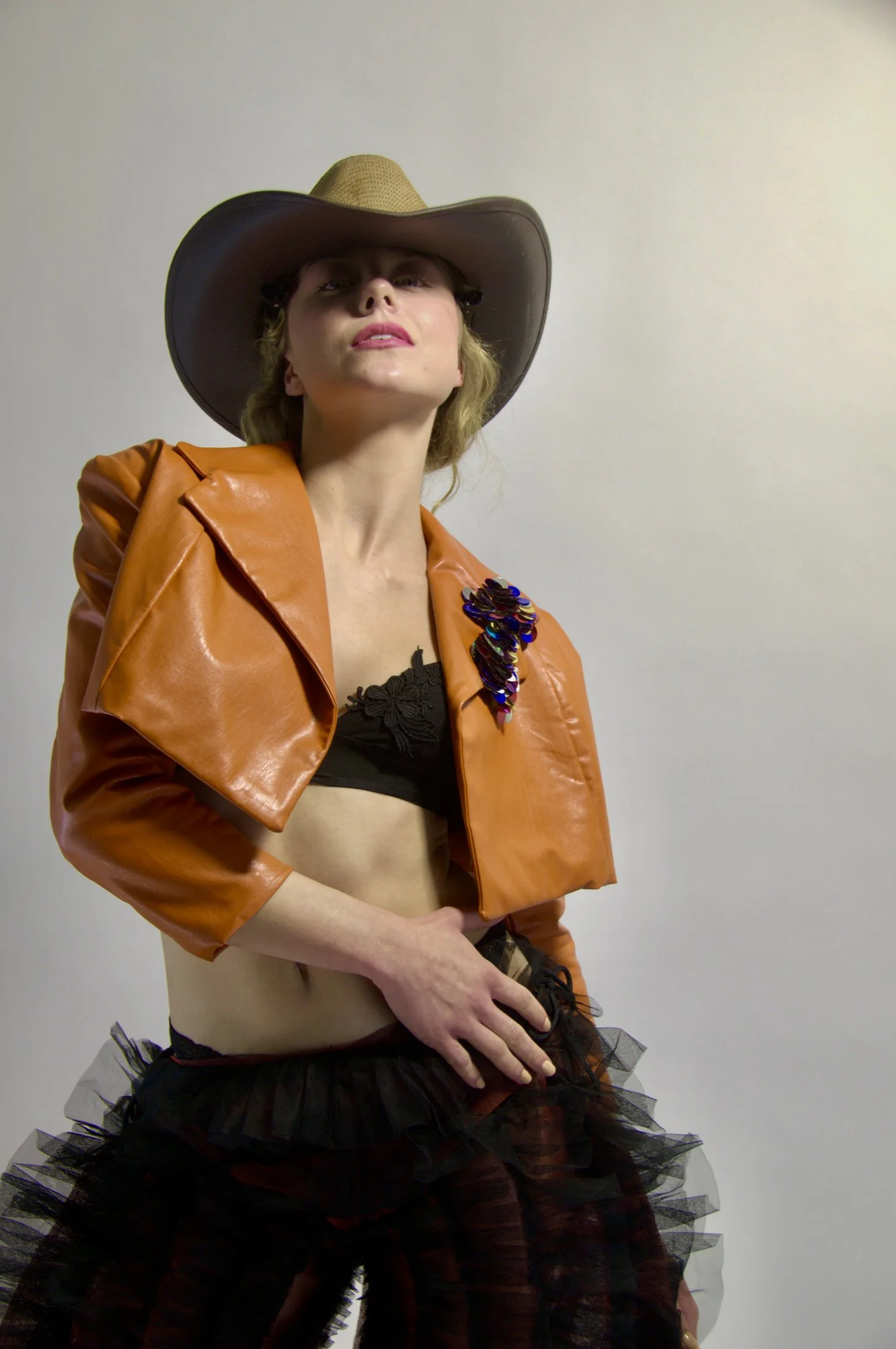A woman wearing a large brown cowboy hat, an orange leather jacket with a sequined embellishment, black lace crop top, gray tutu skirt, and black and orange stockings, standing against a plain background.