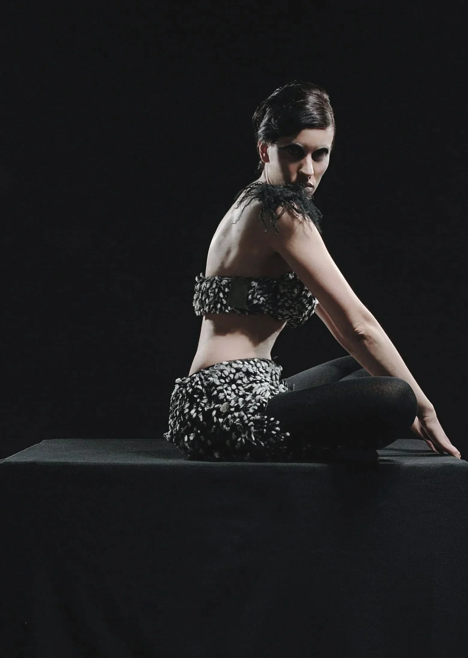 A woman with short dark hair sitting on a black mat against a black background, wearing a textured cloth top and black leggings, with her right arm extended forward and looking directly at the camera.