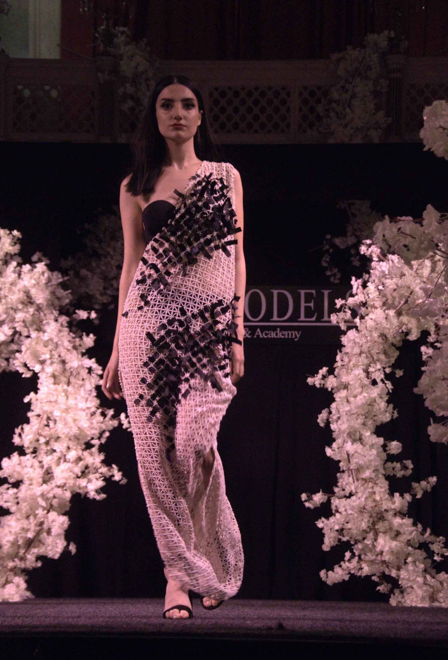 A woman walking on a runway wearing a black and pink multiple layered dress with intricate lattice patterns and black ribbon accents, surrounded by white flowers, at a fashion show event.
