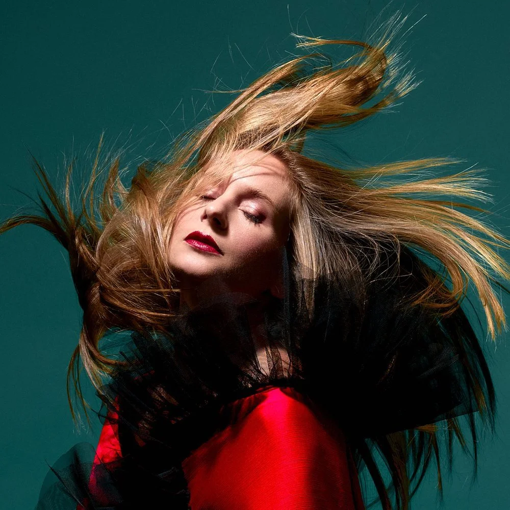A woman with long, flowing blonde hair and red lipstick, wearing a black tulle and red clothing, against a dark teal background.